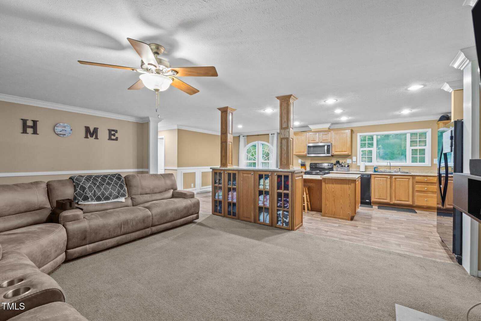 An open living room with a beige sectional sofa, ceiling fan, and carpeted floor leads into a kitchen with wood cabinets, stainless steel appliances, and a central island with built-in shelving.
