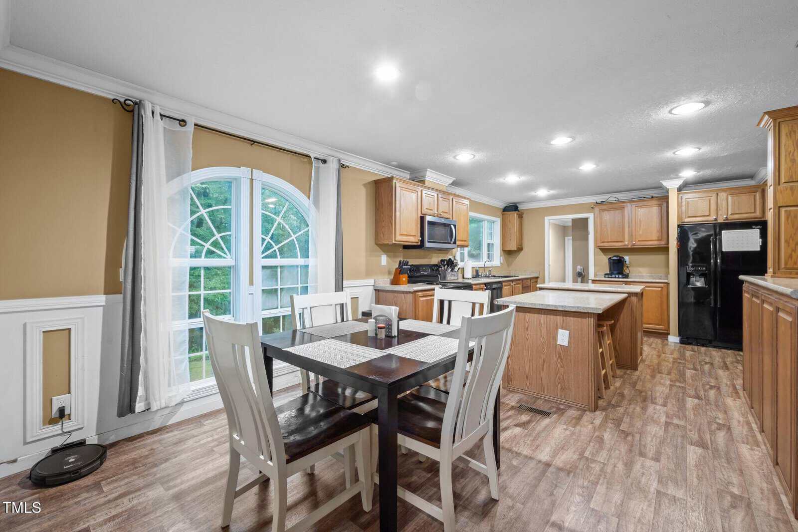 Spacious kitchen with wood cabinets, black appliances, island, dining table for six, arched window with white curtains, tan and white walls, and wood flooring.