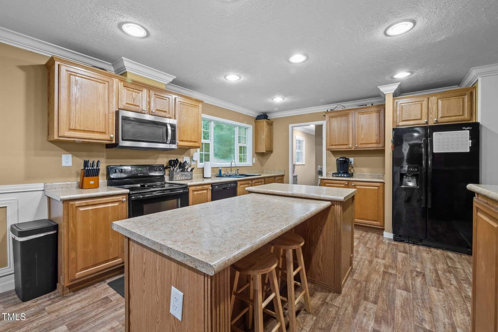 A kitchen with wood cabinets, black appliances, beige countertops, a center island with two stools, and recessed ceiling lights. The room has tan walls, white trim, and a window above the sink.