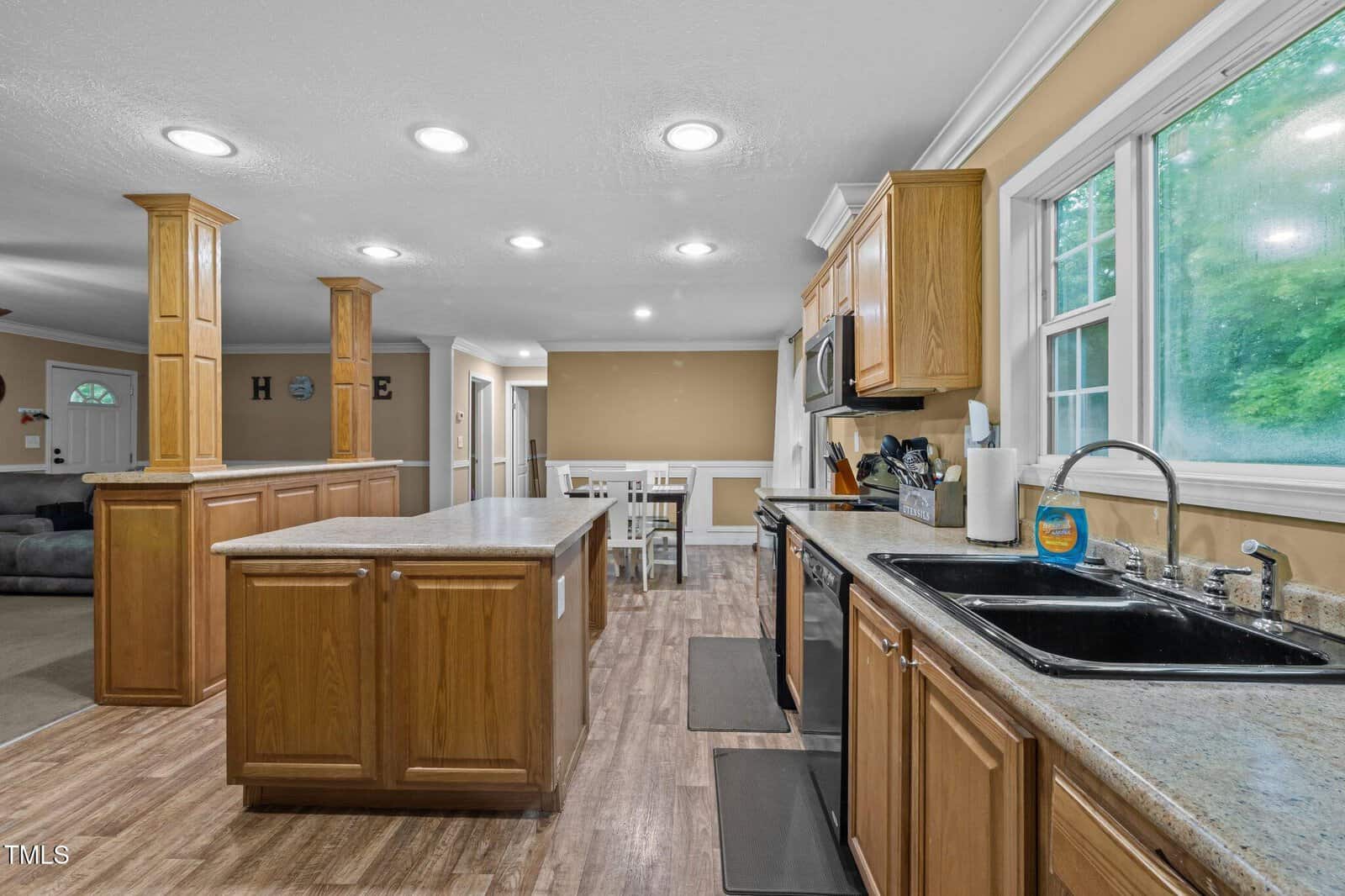 Spacious kitchen with wood cabinets, granite countertops, a central island, double sink, and large windows overlooking greenery. Dining table and living area visible in the background.