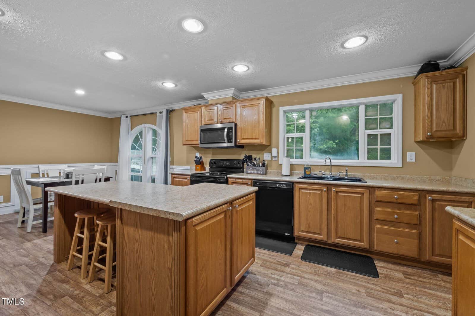 A kitchen with wooden cabinets, a center island with stools, black appliances, and a double sink under a window. Adjacent is a dining area with a table and chairs near glass doors with curtains.