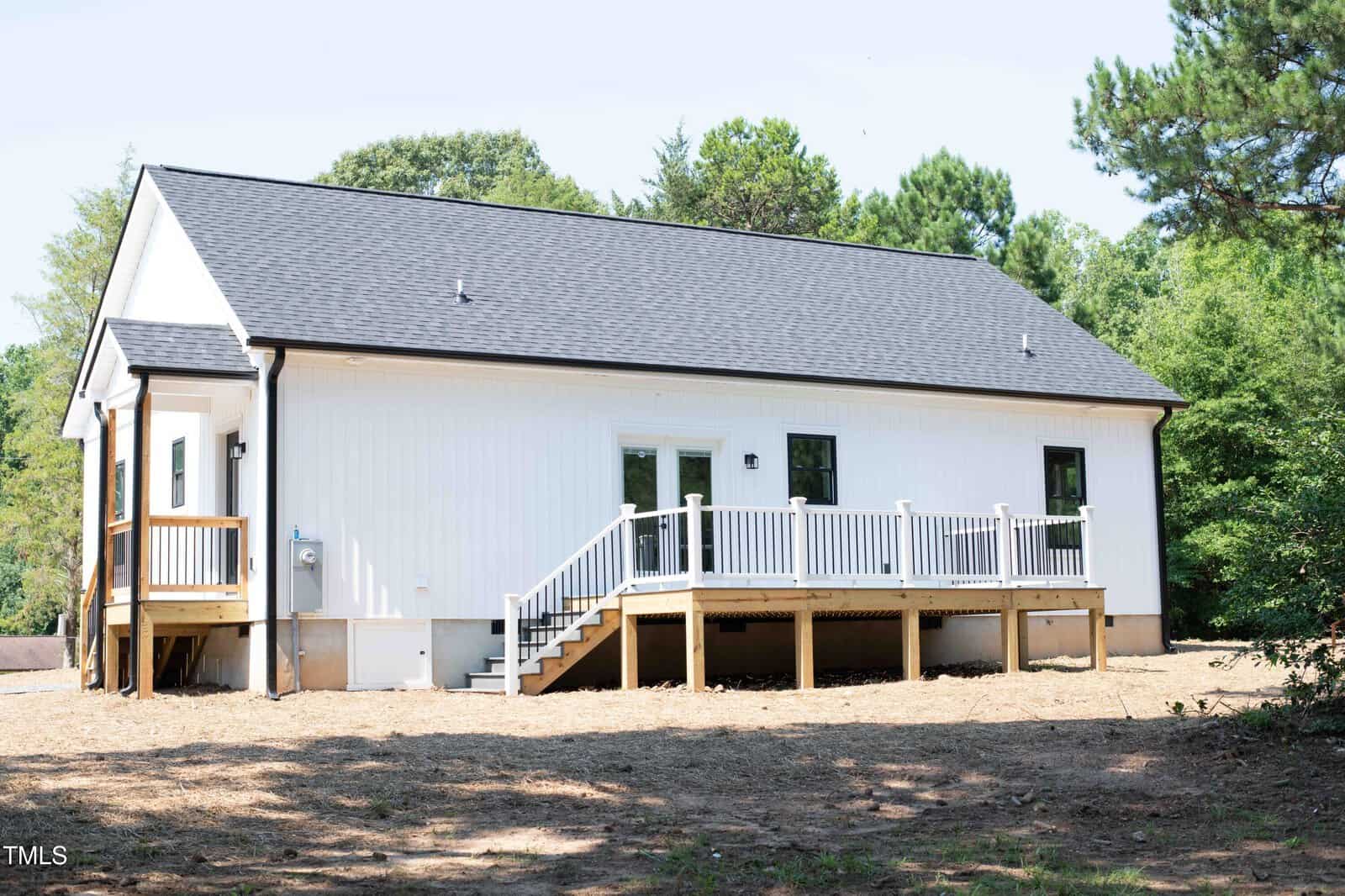 Single-story white house with black roof, small deck and stairs leading to a backyard area. There are several windows and a glass door opening onto the deck, surrounded by trees and natural landscaping.