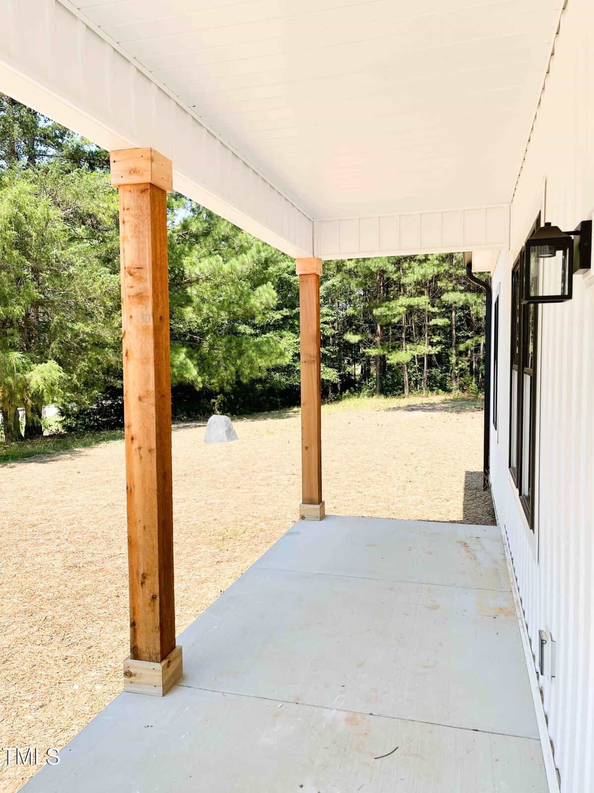 A covered porch with a concrete floor, wooden support columns, and a white ceiling. The porch overlooks a grassy yard with trees in the background. The exterior wall of the house is white with black-trimmed windows.