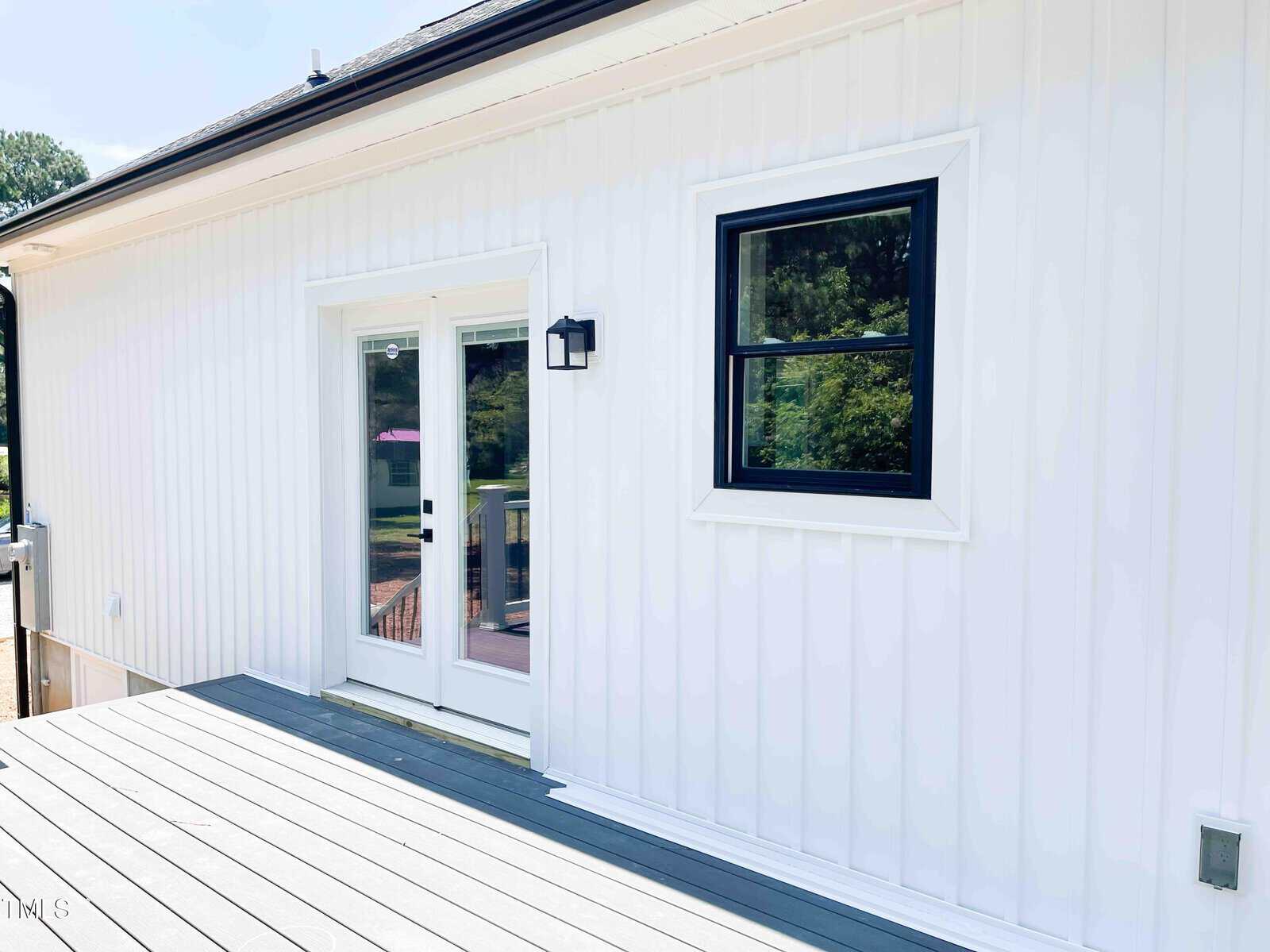 A white exterior wall with vertical siding features a black-framed window and glass double doors. A black outdoor wall light is mounted beside the doors. The deck below is light gray. Trees are visible in the reflection.