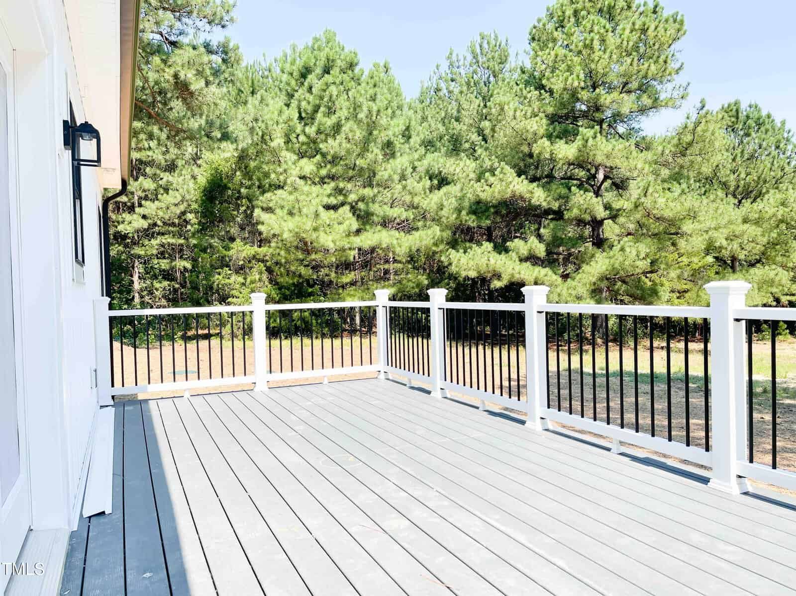 A spacious wooden deck with white and black railing is attached to a house, overlooking a grassy area and dense green trees under a clear sky.