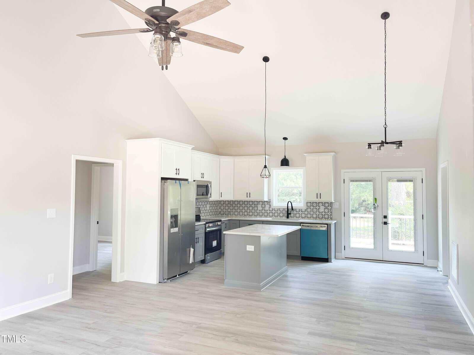Modern kitchen with light wood floors, white cabinets, stainless steel appliances, pendant lights, patterned backsplash, small island, and French doors leading outside. Ceiling fan and high ceilings are visible.