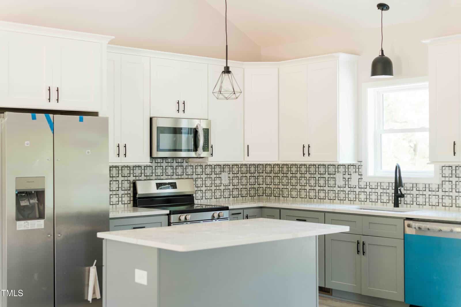 Bright kitchen with white upper cabinets, gray lower cabinets, a kitchen island, stainless steel appliances, black and white tile backsplash, pendant lights, and a window above the sink.