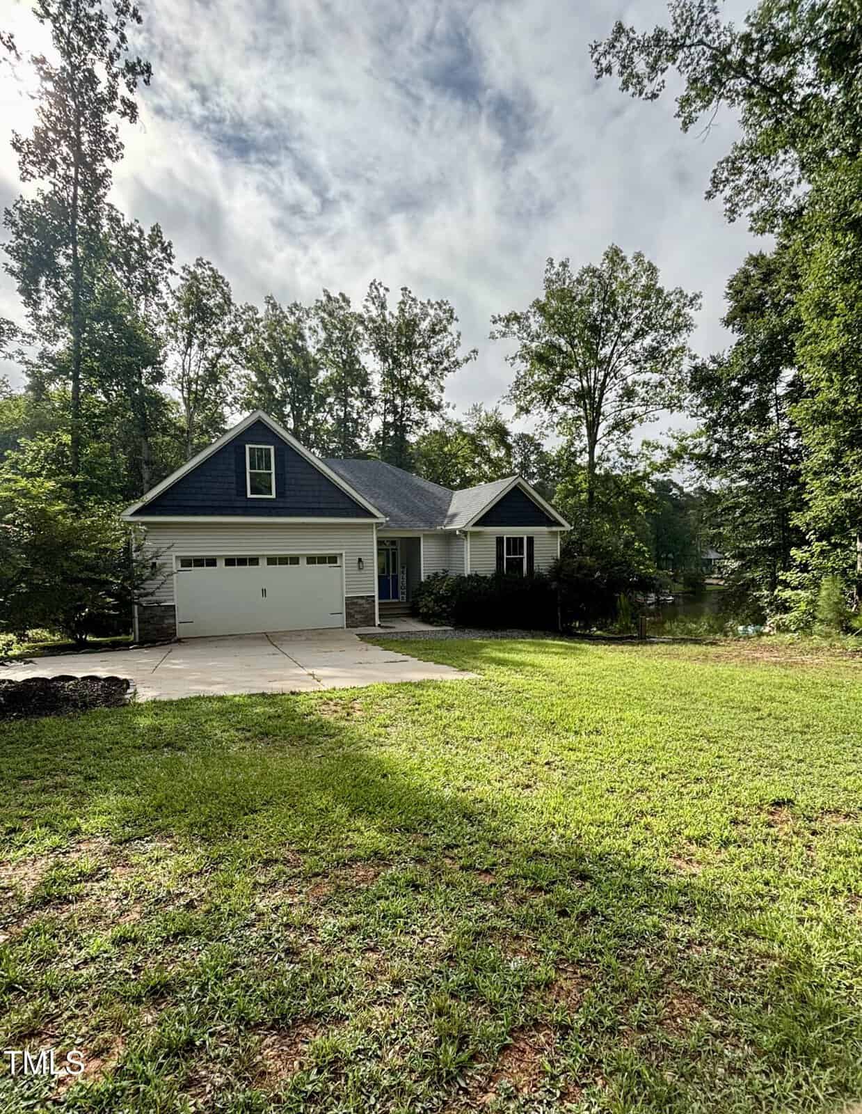 A single-story house with blue siding and white trim sits among tall trees. A wide driveway leads to a double garage, and a well-kept lawn fills the foreground under a partly cloudy sky.