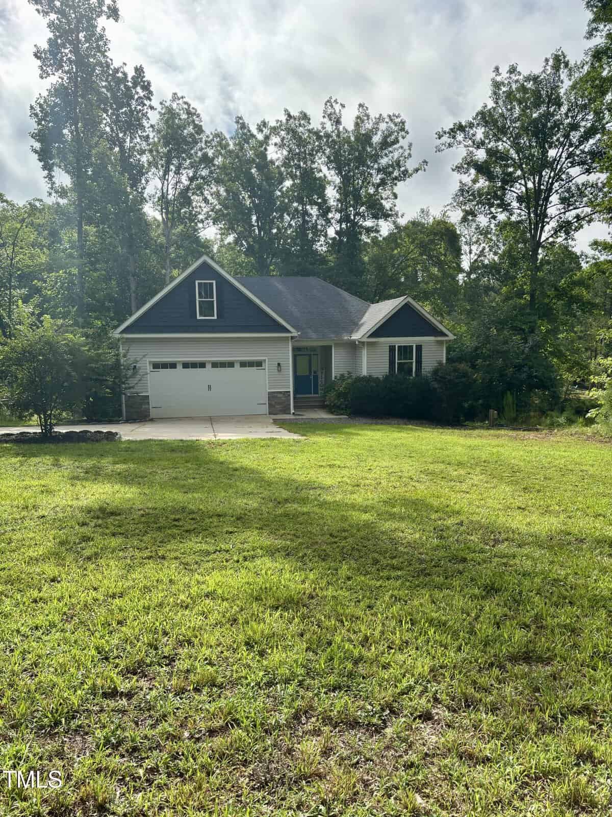 A small, dark gray house with a white garage and front porch sits amid a large green lawn, surrounded by tall trees and dense foliage under a cloudy sky.