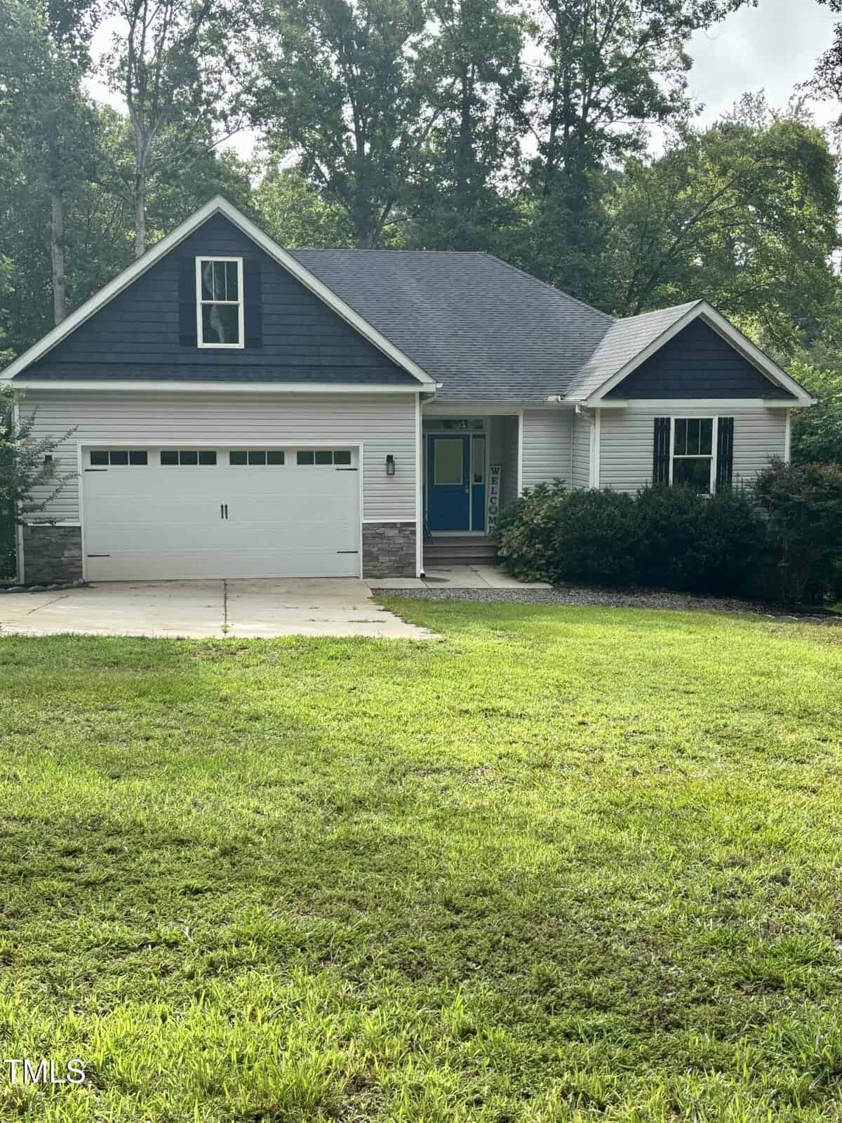 Single-story house with dark blue and gray siding, a two-car garage, a front porch with a blue door, and a sloped roof. The house is surrounded by green grass, trees, and bushes.