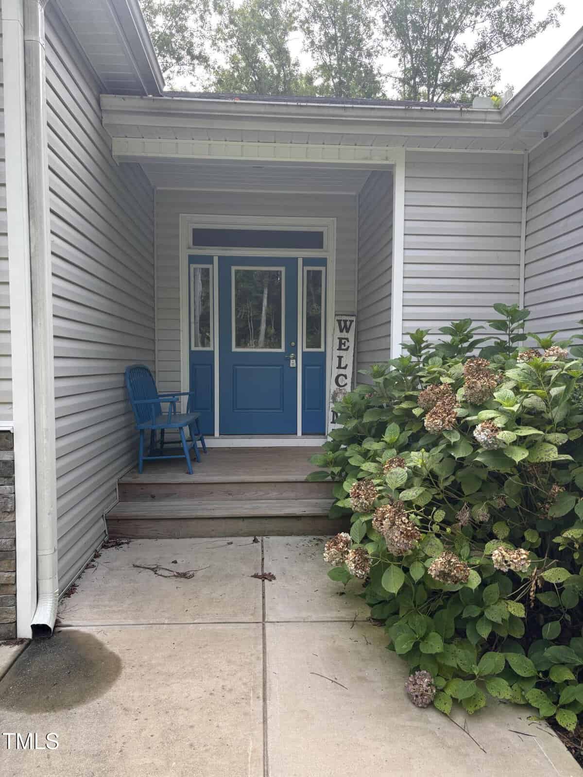 A front porch with a blue door, blue chair, “Welcome” sign, and a bush with faded hydrangea flowers beside a concrete walkway. The house has light gray siding and white trim.