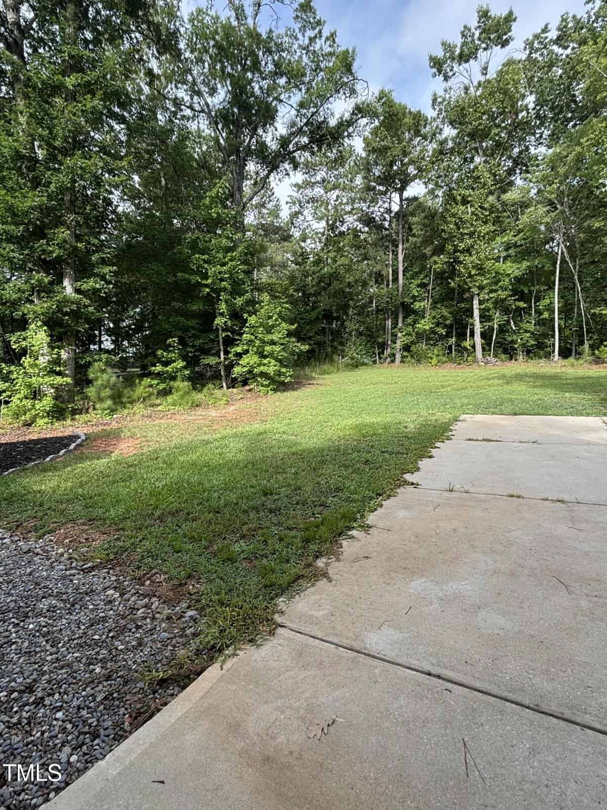 A concrete patio leads to a grassy lawn bordered by a mulched area and tall, leafy trees under a partly cloudy sky.