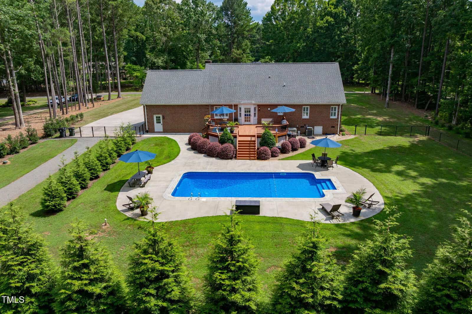 A brick house with a large back deck overlooks a rectangular swimming pool surrounded by a concrete patio. Lounge chairs and umbrellas are set up around the pool, and the yard is bordered by tall trees and greenery.