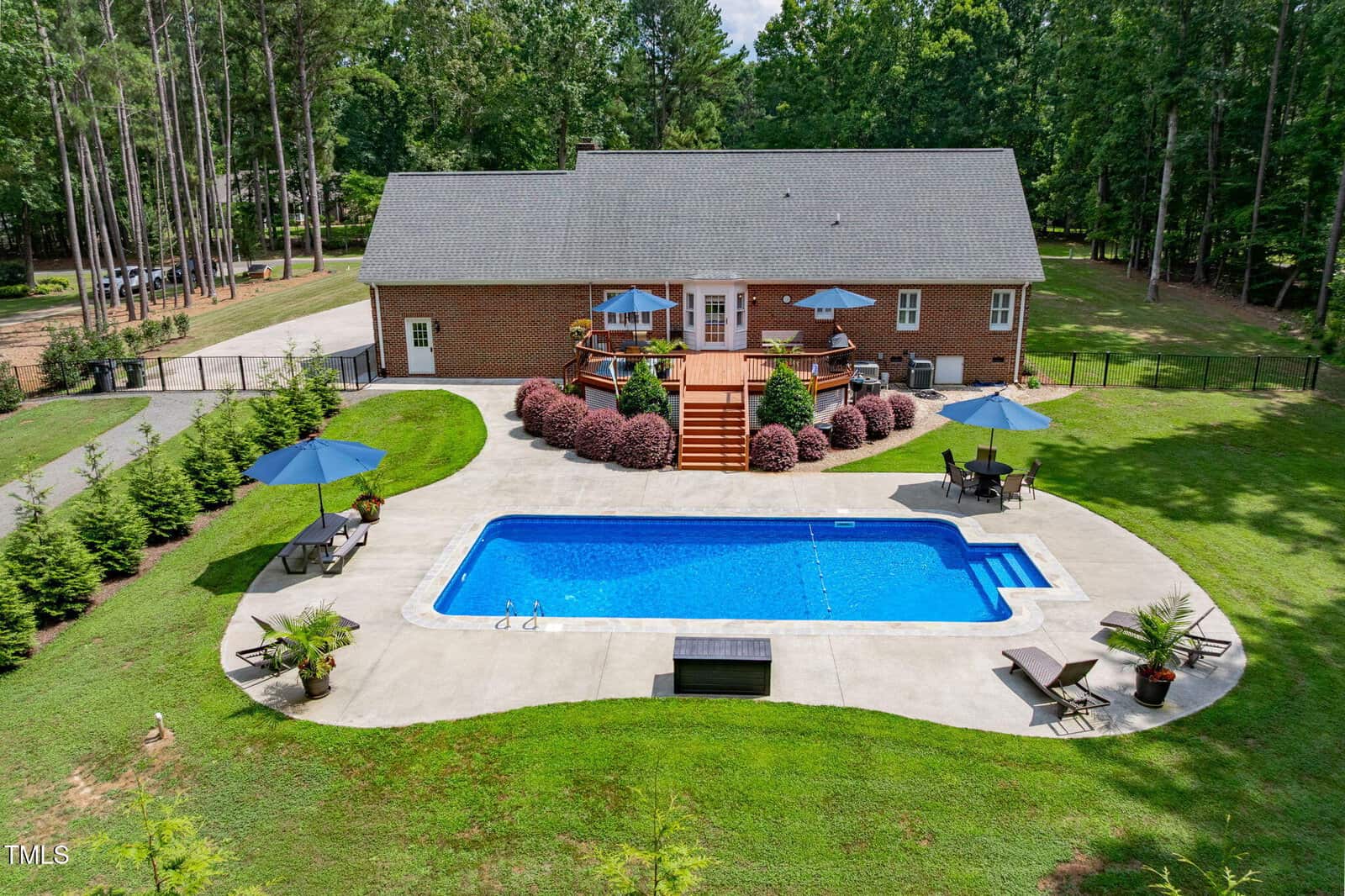 A large brick house with a spacious wooden deck overlooks a rectangular in-ground swimming pool. The pool area is surrounded by patio furniture, umbrellas, and landscaped greenery, with a fenced yard and tall trees in the background.