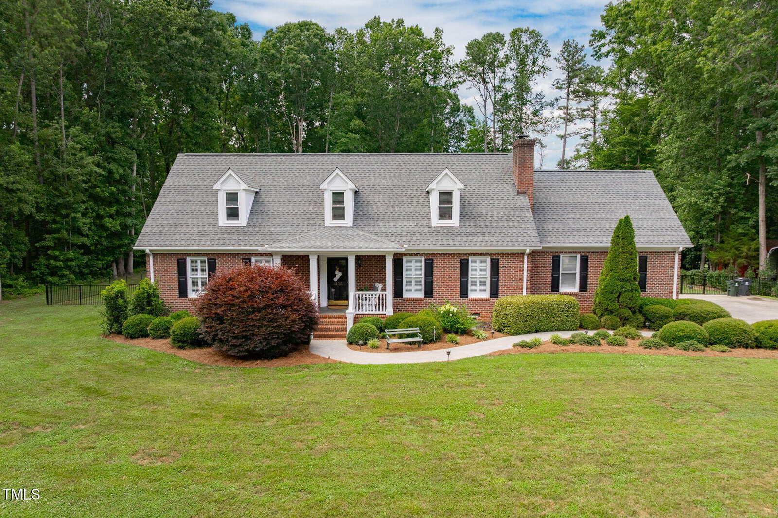 A brick one-and-a-half-story house with dormer windows, a covered front porch, and a manicured lawn. The house is surrounded by trees and landscaped bushes, with a driveway on the right side.