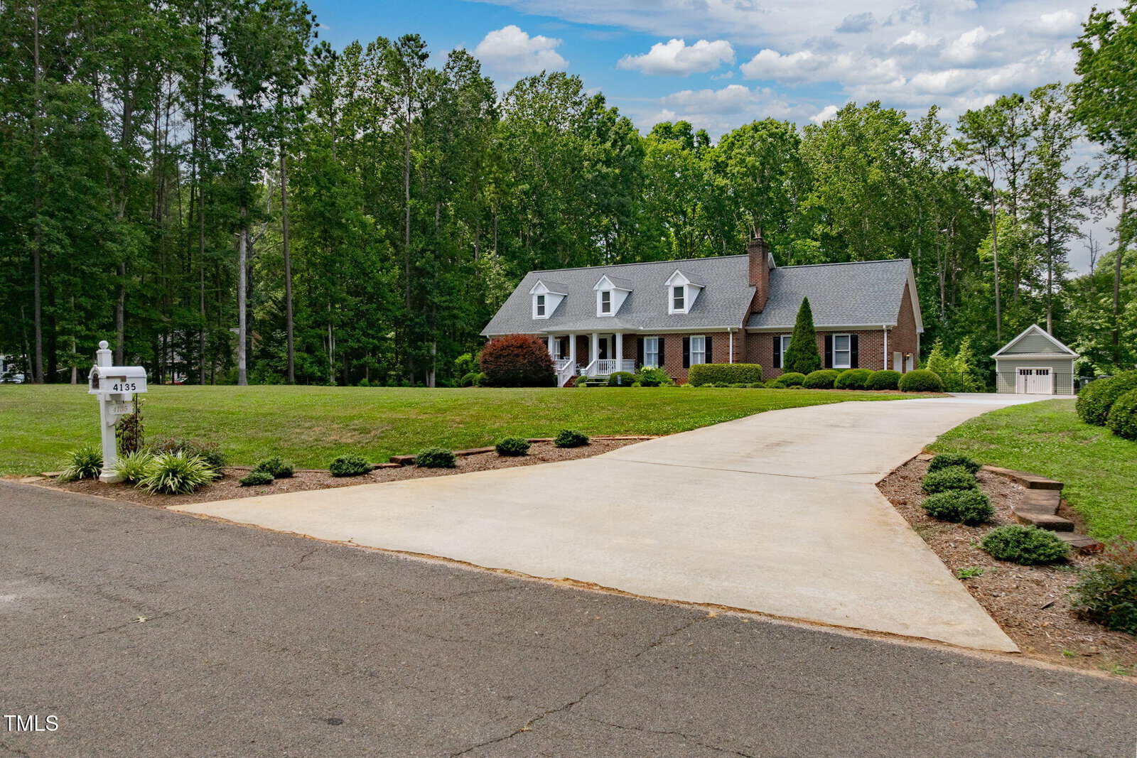 A brick house with white trim, dormer windows, and a large front lawn sits at the end of a long driveway, surrounded by trees. A detached garage and white mailbox are also visible.