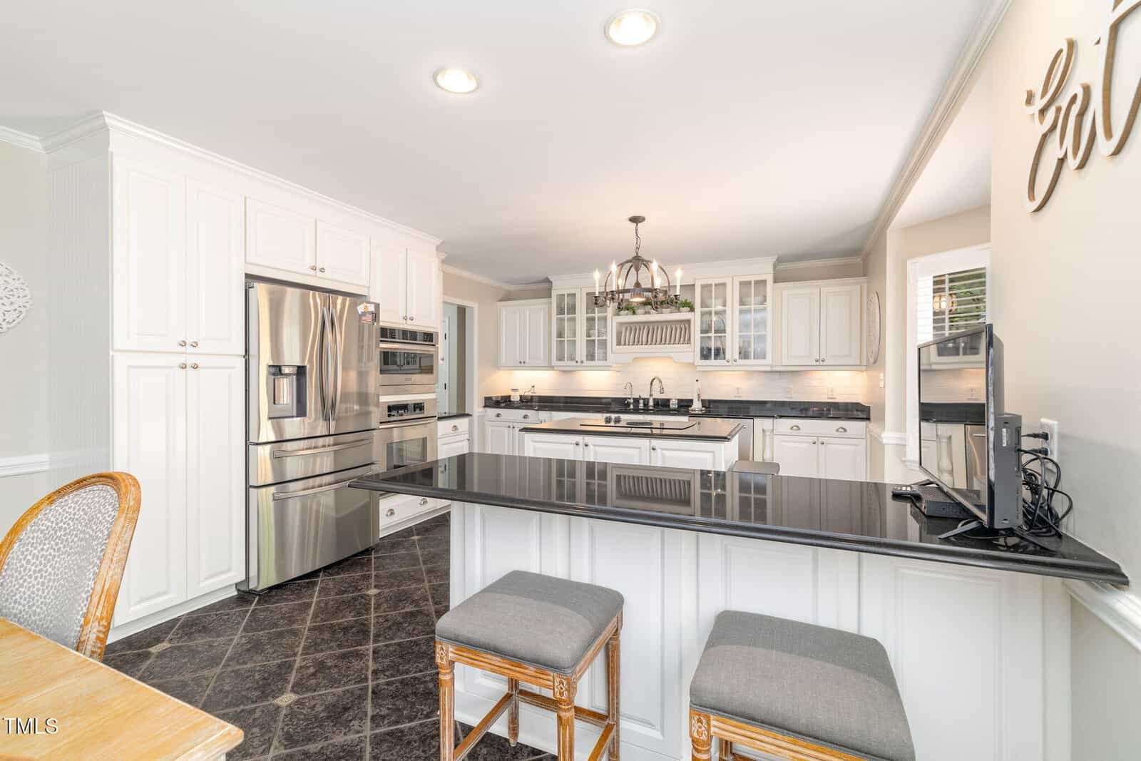 Bright, modern kitchen with white cabinets, stainless steel appliances, black countertops, a large island with two stools, and a chandelier overhead. The space is clean and inviting with plenty of natural light.