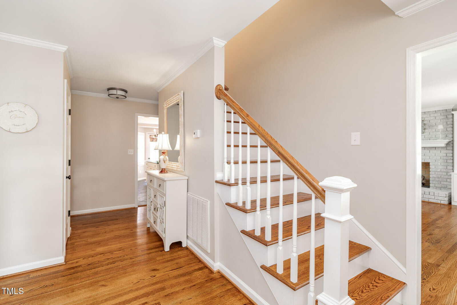 A hallway with light walls, hardwood floors, a white staircase with wooden handrail, a white dresser with mirror, and a glimpse of a living room with a brick fireplace.