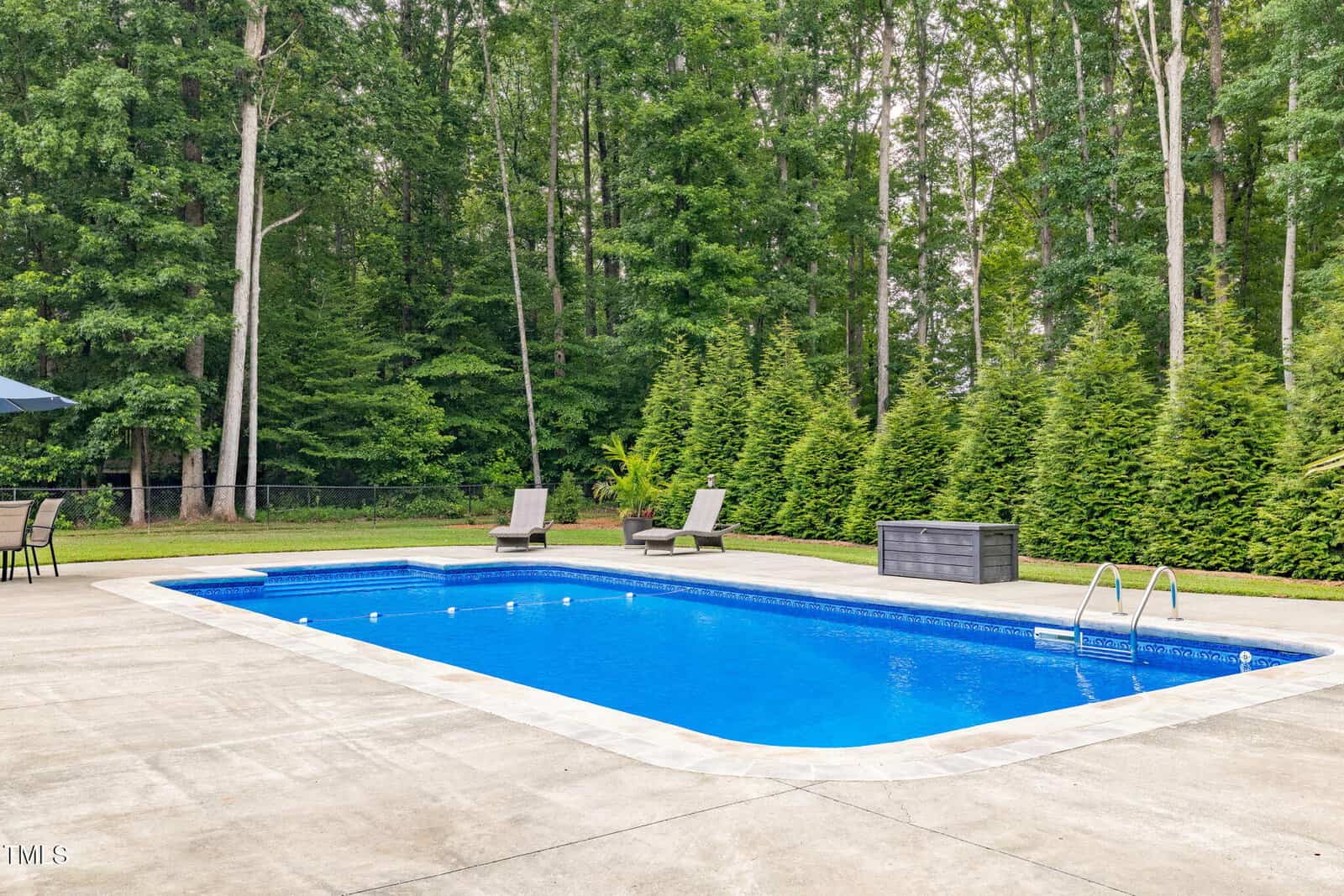 Rectangular inground swimming pool with steps, surrounded by a concrete patio. Two lounge chairs and a storage box are beside the pool, with a backdrop of tall trees and shrubs. Outdoor seating is visible on the left.
