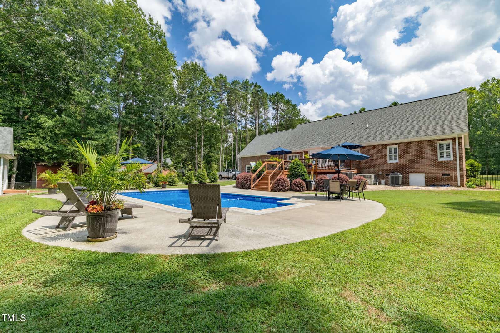 A backyard with a rectangular in-ground pool surrounded by a concrete patio, lounge chairs, potted plants, and umbrellas. A large brick house with a deck and stairs is in the background, with trees lining the yard.