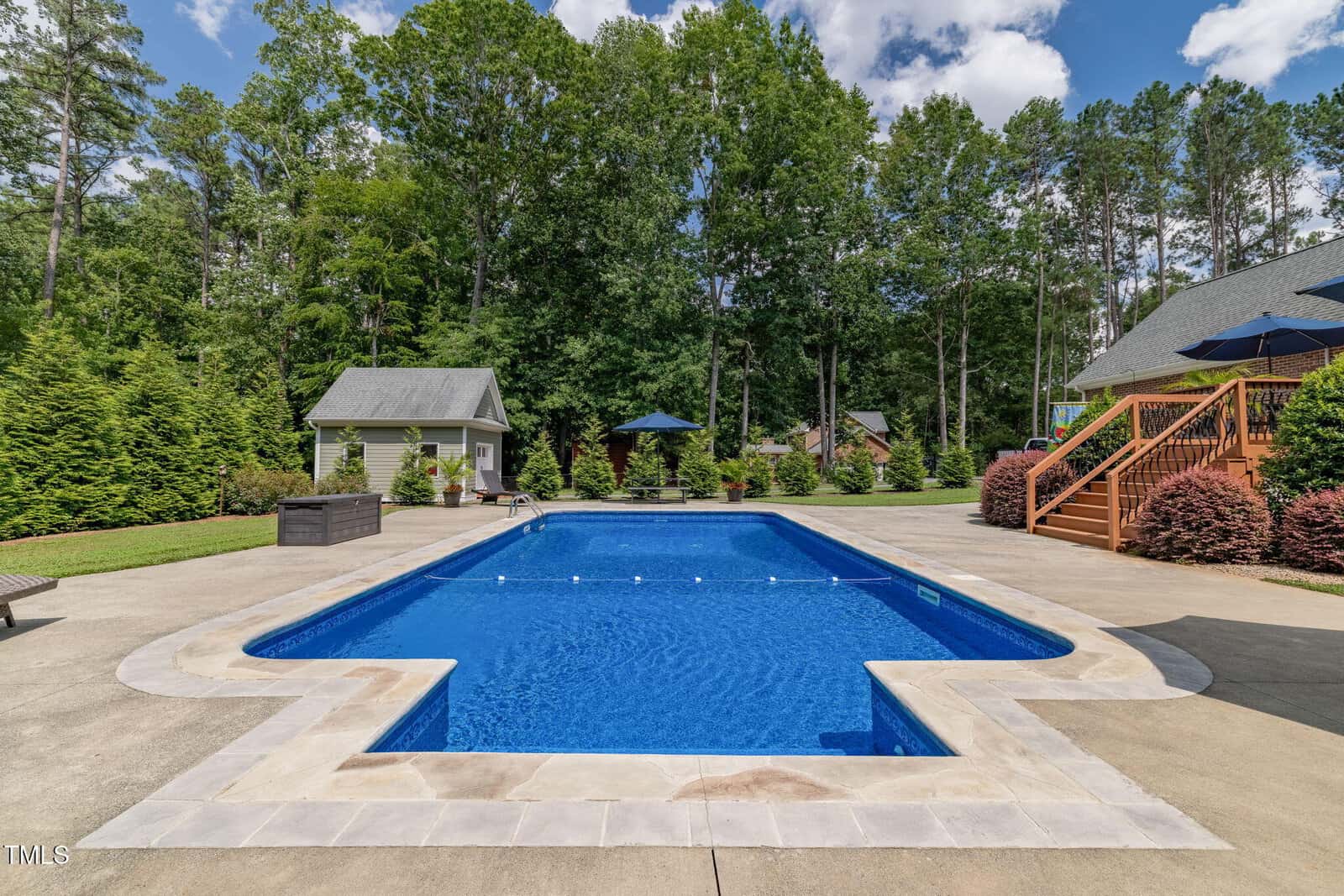 A rectangular in-ground swimming pool with a concrete deck, surrounded by trees and shrubs. There are lounge chairs, umbrellas, a small building, and a wooden staircase leading to a raised deck on the right.