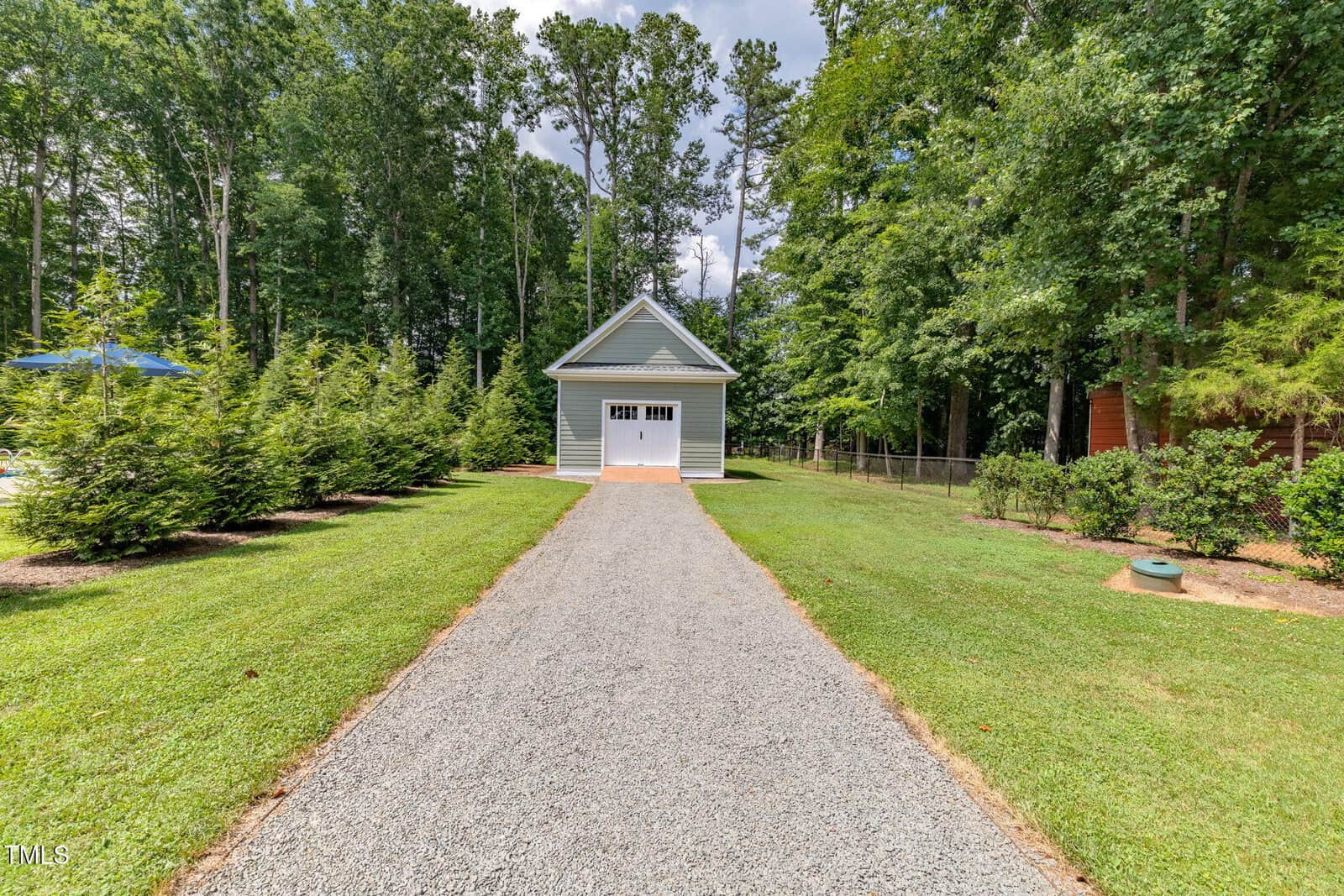 A gravel driveway leads to a small gray garage with white trim and double doors, surrounded by green grass, trees, and landscaping on a sunny day.