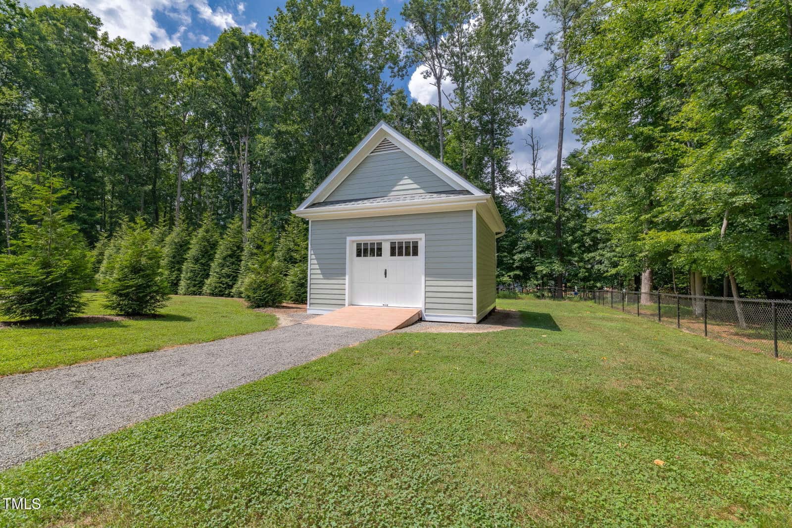 A small, gray detached garage with white trim and a single garage door sits on a gravel path surrounded by grass and trees, with a chain-link fence visible to the right.