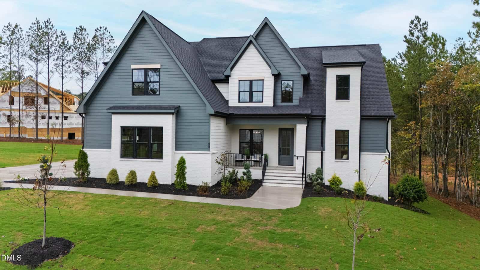 Modern two-story house with white brick and gray siding, black-framed windows, front porch with chairs, neatly landscaped front yard, and trees in the background.