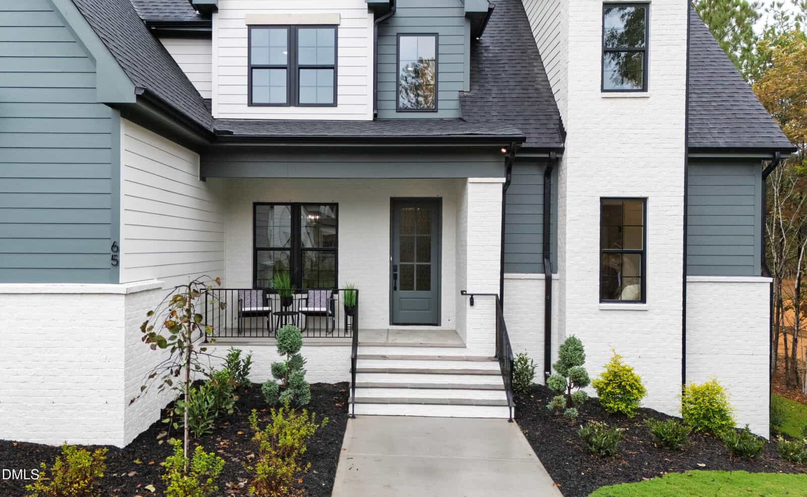 Modern two-story house with grey and white exterior, black trim, large windows, small porch with two chairs, steps leading to a glass front door, and neatly landscaped front yard.