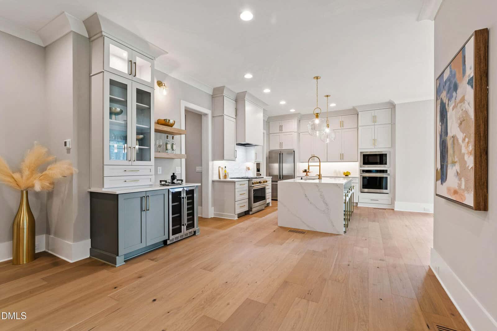 A modern kitchen with light wood floors, white cabinetry, a large marble island, stainless steel appliances, and gold accents. Glass cabinets, a wine fridge, and abstract art decorate the space.
