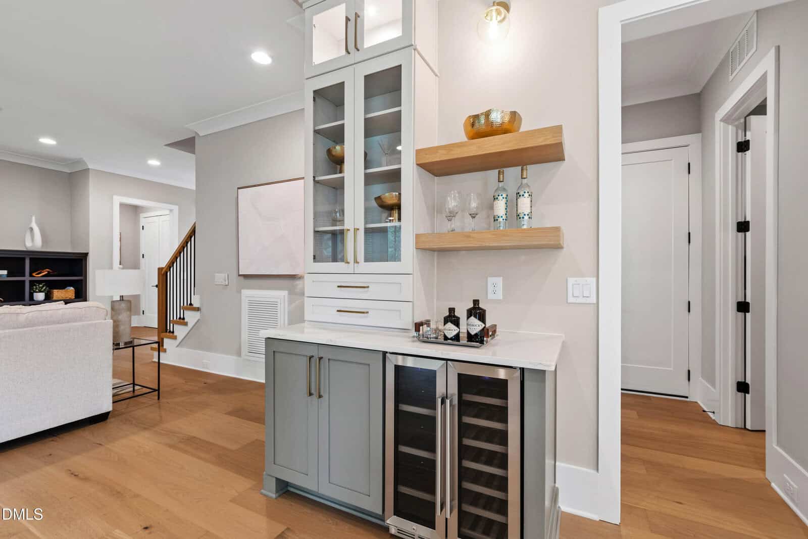 Modern home bar area with glass-front cabinets, open wood shelves with decor, a marble countertop, a wine fridge, and a tray with bottles and glasses. Light wood flooring and neutral walls complete the space.