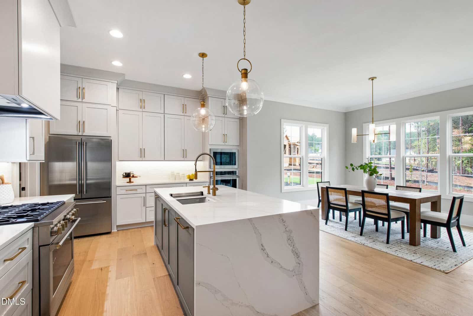 Modern kitchen with a marble island, stainless steel appliances, pendant lights, white cabinets, and a nearby dining area with large windows providing natural light and a view of a backyard playground.