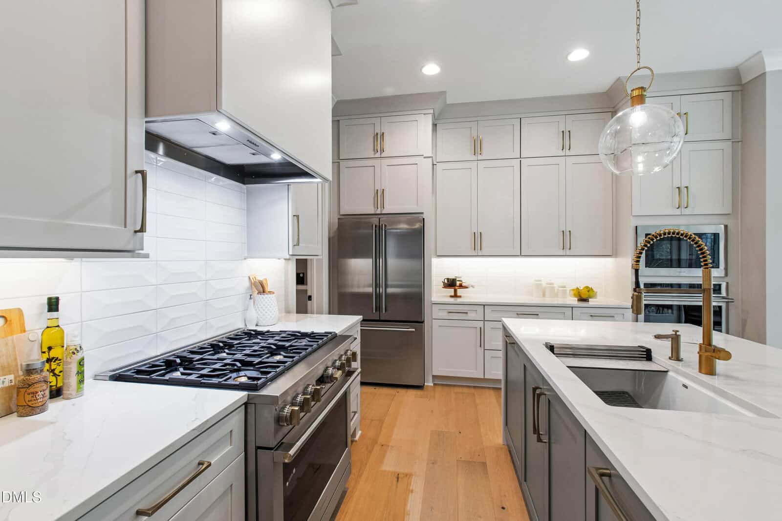 Modern kitchen with white cabinets, stainless steel appliances, a large refrigerator, gas stove, built-in oven, white countertops, gold faucet, under-cabinet lighting, and wooden floors.