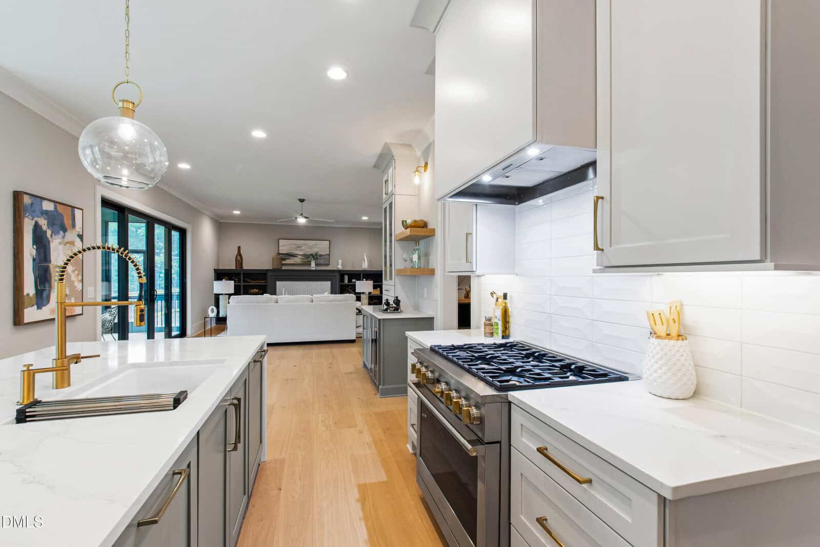 Modern kitchen with white cabinets, gold fixtures, a large gas stove, white countertops, a farmhouse sink, and light wood floors. The space opens into a living room with a fireplace and large windows.