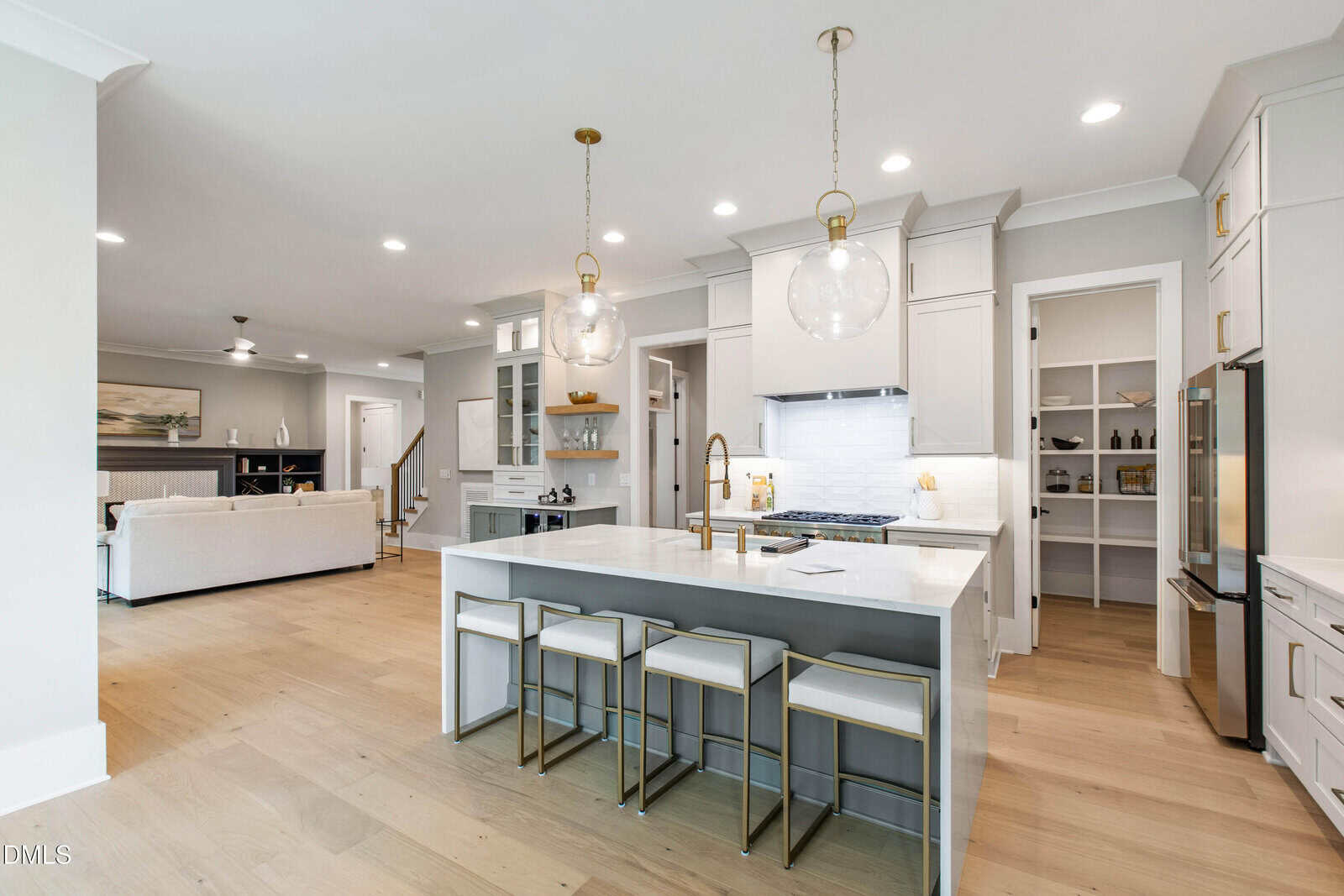 Modern kitchen with white cabinets, marble island with four stools, gold pendant lights, and stainless steel appliances; open layout leads to a living area with neutral decor and wooden floors throughout.