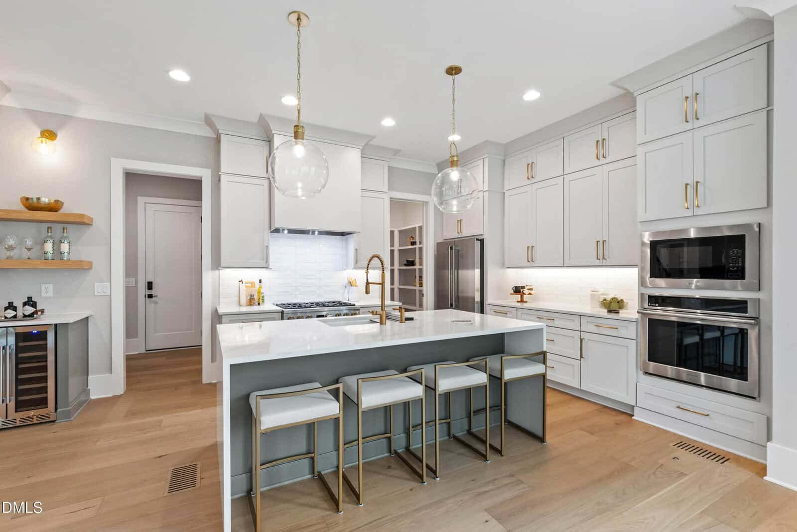 Modern kitchen with white cabinets, stainless steel appliances, a large island with four stools, gold accents, pendant lights, light wood floors, and a bar area with open shelves and wine fridge.