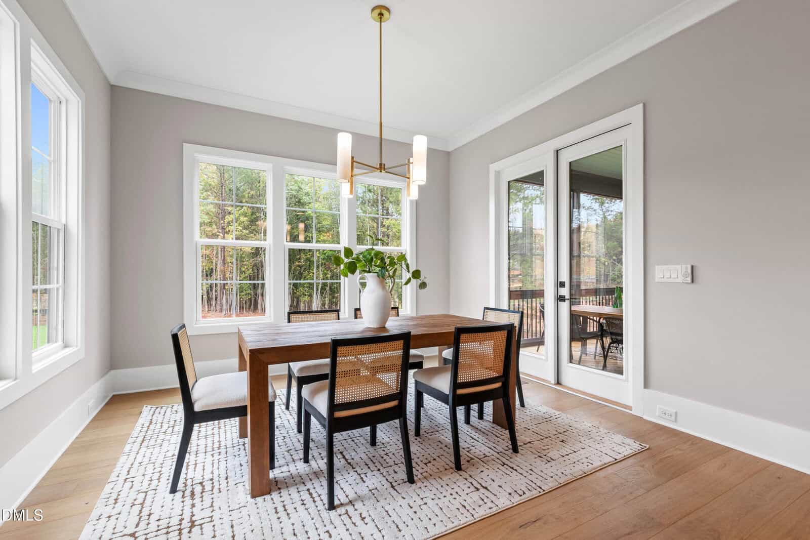 Modern dining room with a wooden table, six black and white chairs, a white rug, a large window, glass doors, light gray walls, wood floors, and a contemporary chandelier overhead. A vase with greenery sits on the table.