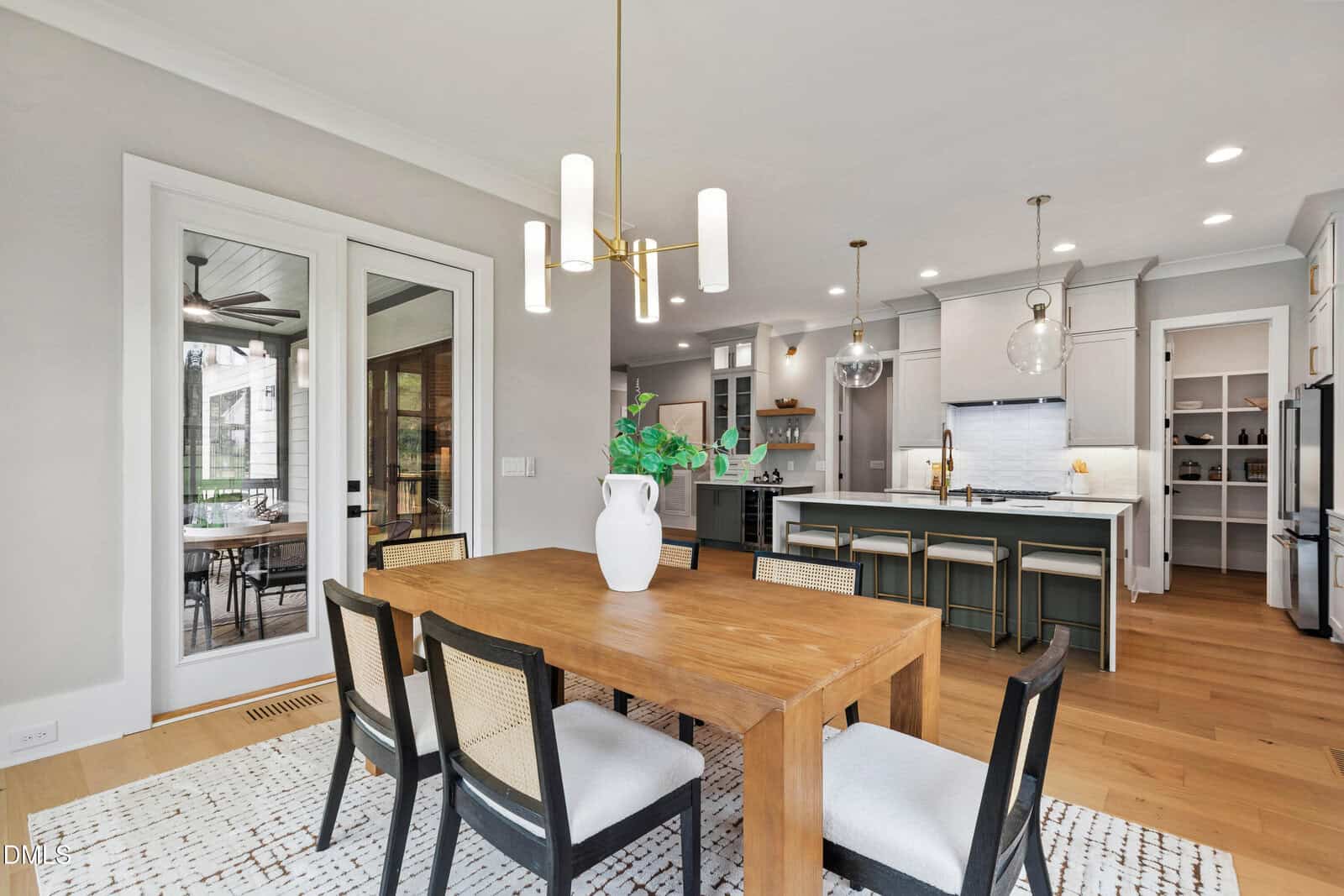Modern dining room with wooden table and six chairs, white vase centerpiece, light fixture above, open kitchen in background with island seating, pendant lights, and stainless steel appliances. Glass doors lead to another room.