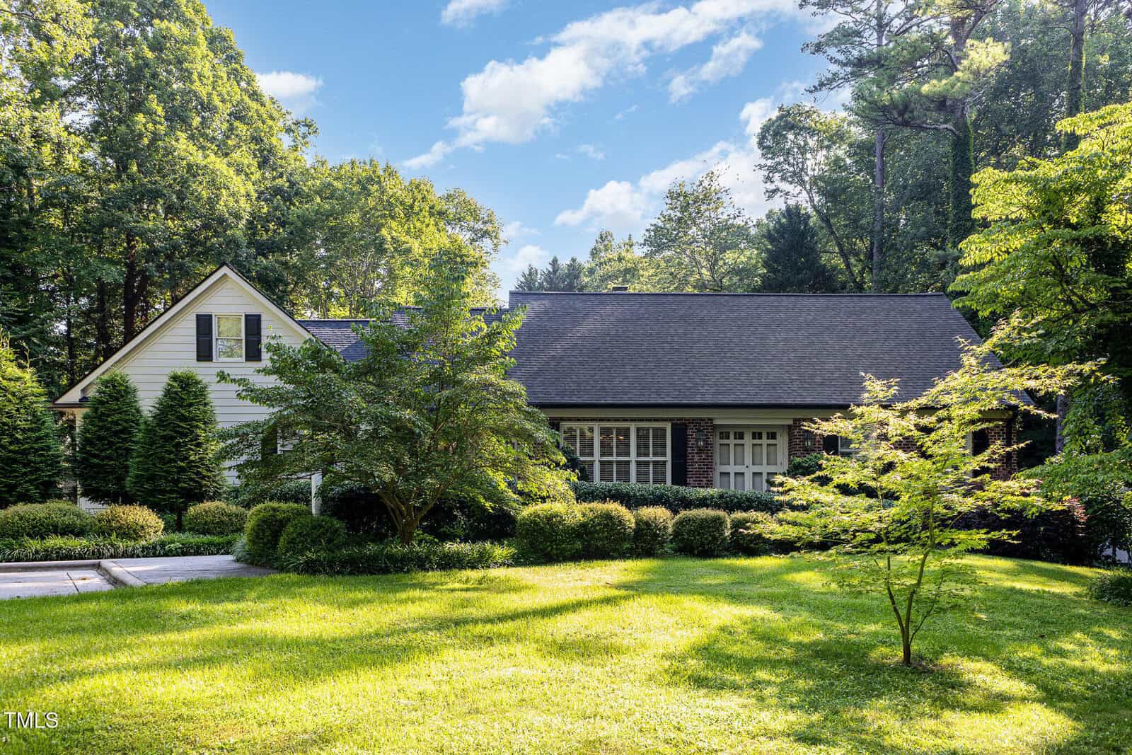 Single-story house with dark roof and white siding, partially obscured by trees and shrubs, situated on a well-maintained lawn surrounded by tall trees under a blue sky with scattered clouds.