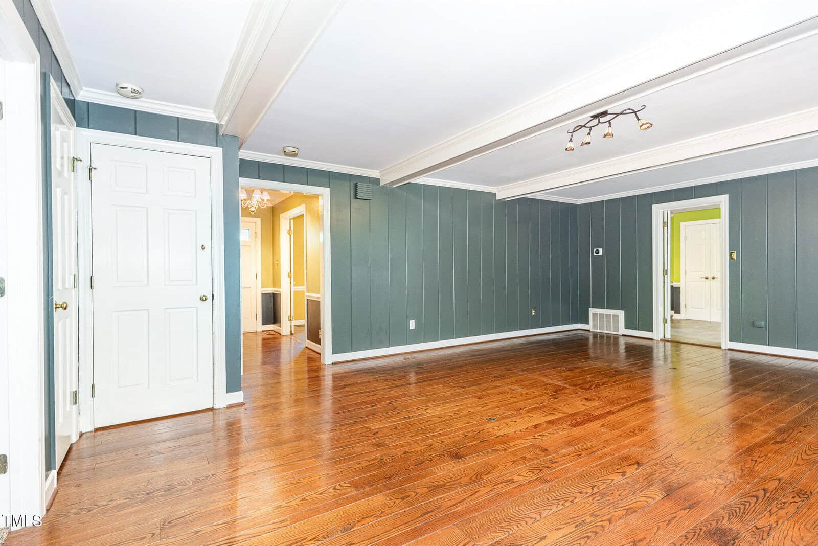 A spacious empty room with green paneled walls, hardwood floors, white trim, ceiling beams, and multiple doorways leading to adjoining rooms. The ceiling has a modern light fixture.