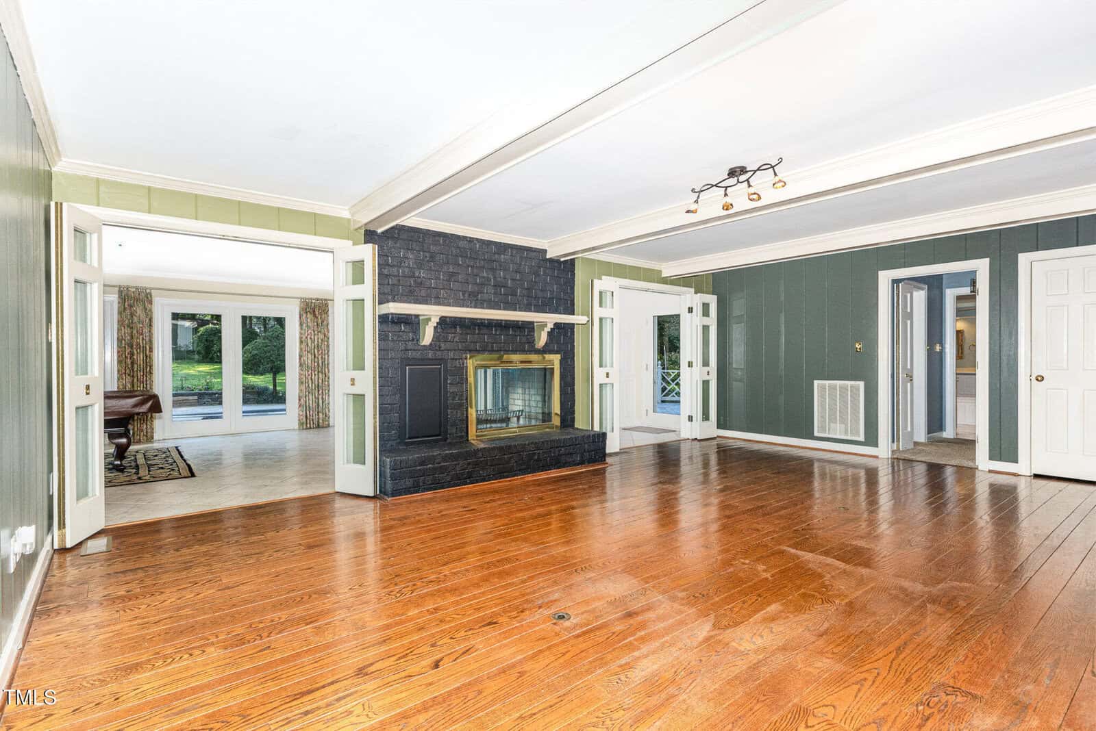 A spacious living room with dark green walls, wood flooring, a black brick fireplace with a brass-trimmed glass door, large windows, and open doorways leading to adjacent rooms and a view of a backyard.