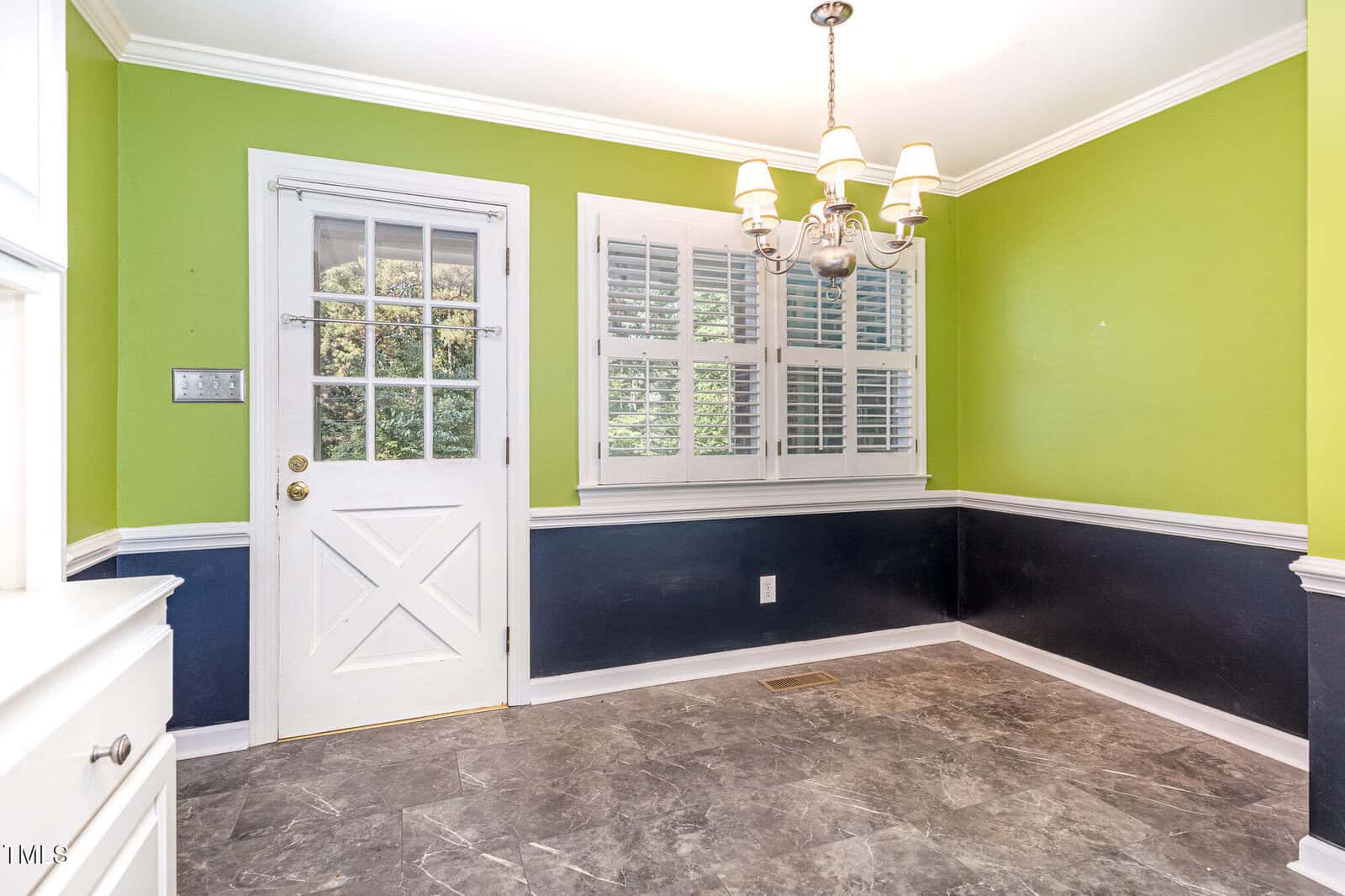 Bright room with green and navy walls, white trim, a window with shutters, a white door, simple chandelier, and gray tiled floor.