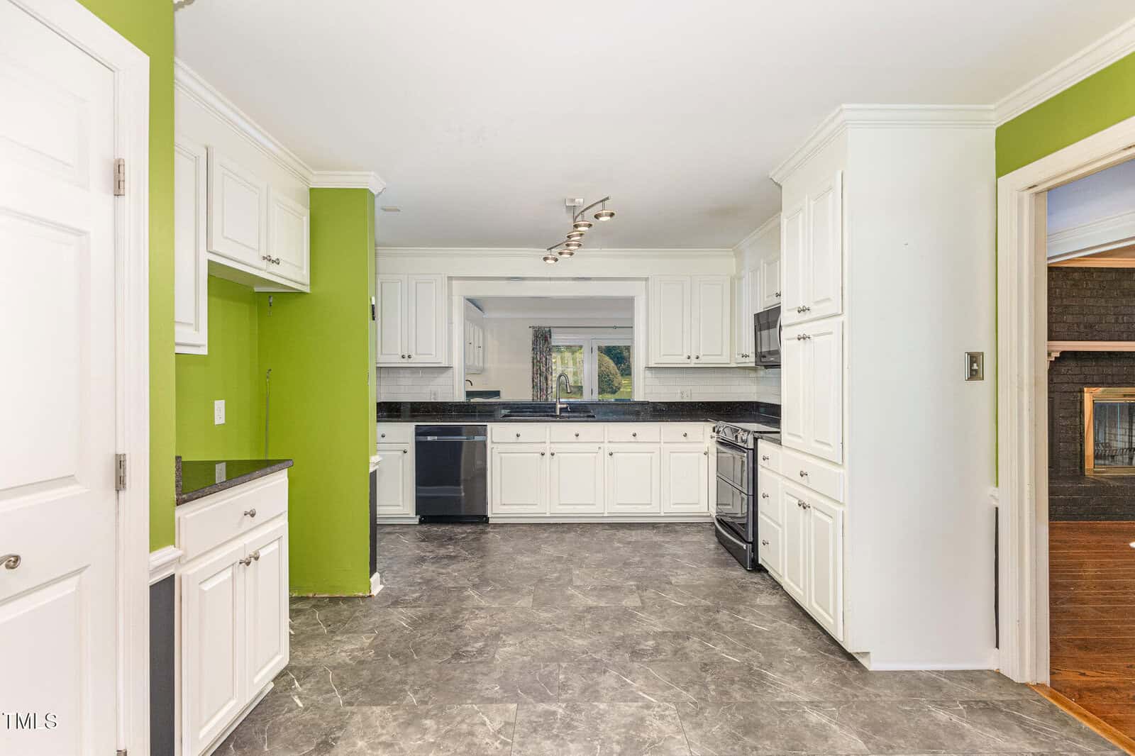 Modern kitchen with white cabinets, black appliances, gray tile floor, green accent walls, and a view into a room with wood floors and a fireplace. Large window above the sink provides natural light.