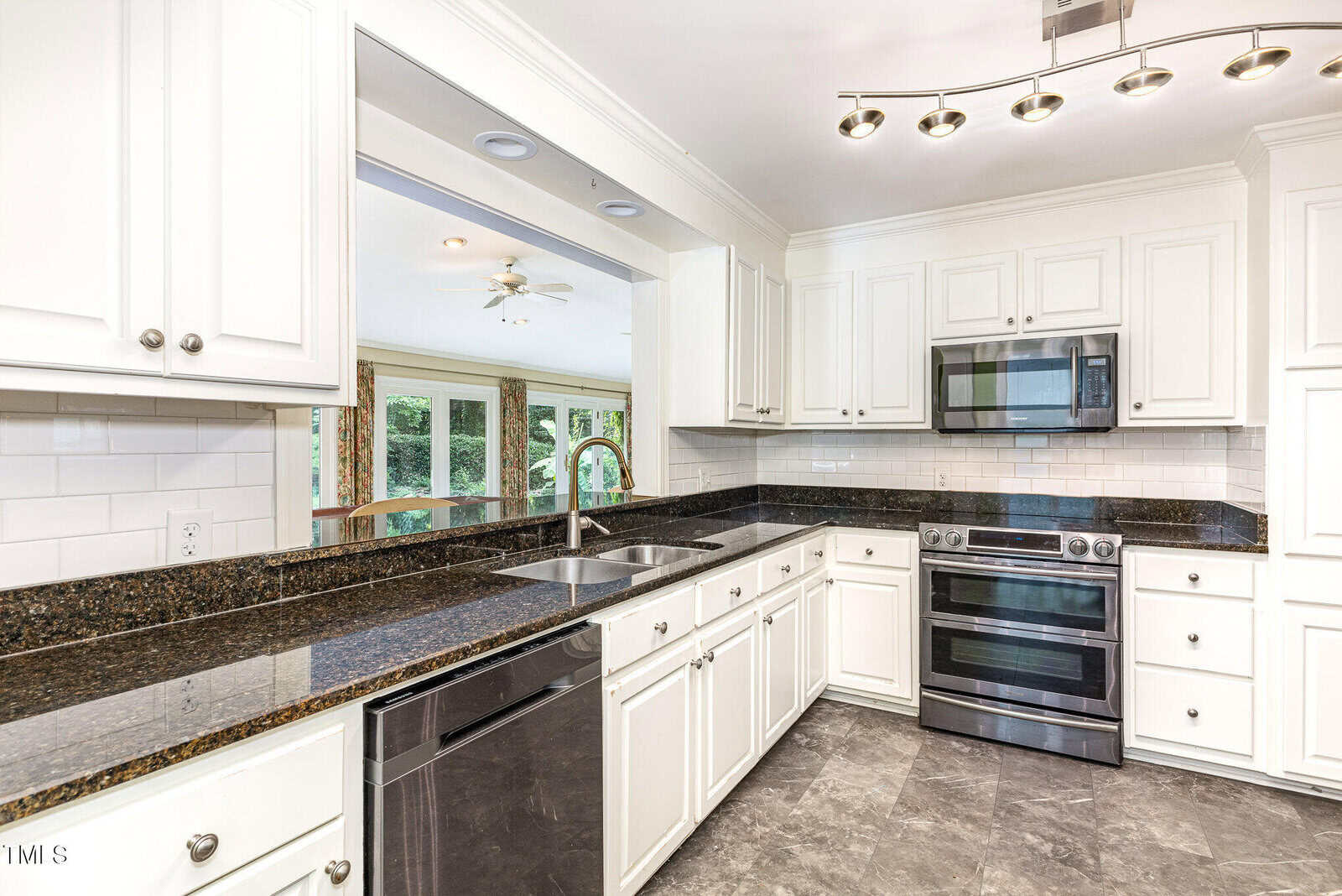 Modern kitchen with white cabinets, dark granite countertops, stainless steel appliances, a double sink, tile backsplash, and gray tile flooring. Large pass-through window opens to a bright living area with large windows.