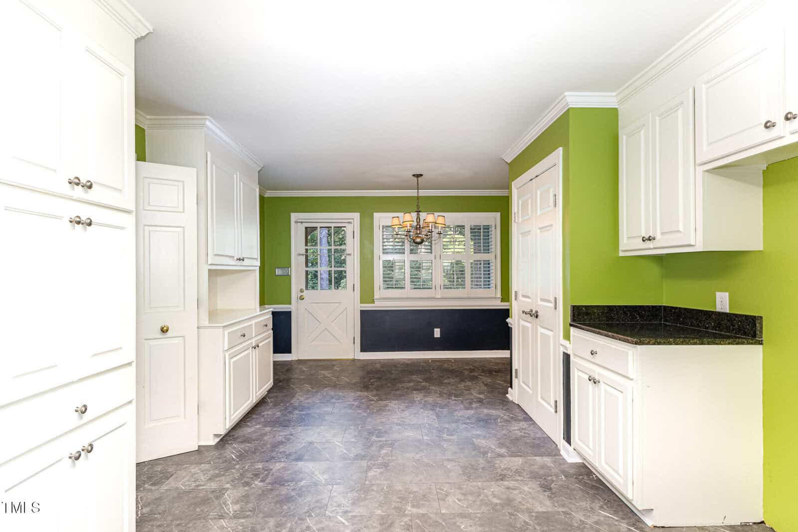A kitchen with green and white walls, white cabinets, black countertops, gray tile flooring, and a chandelier above a dining area near a Dutch door and windows with white shutters.