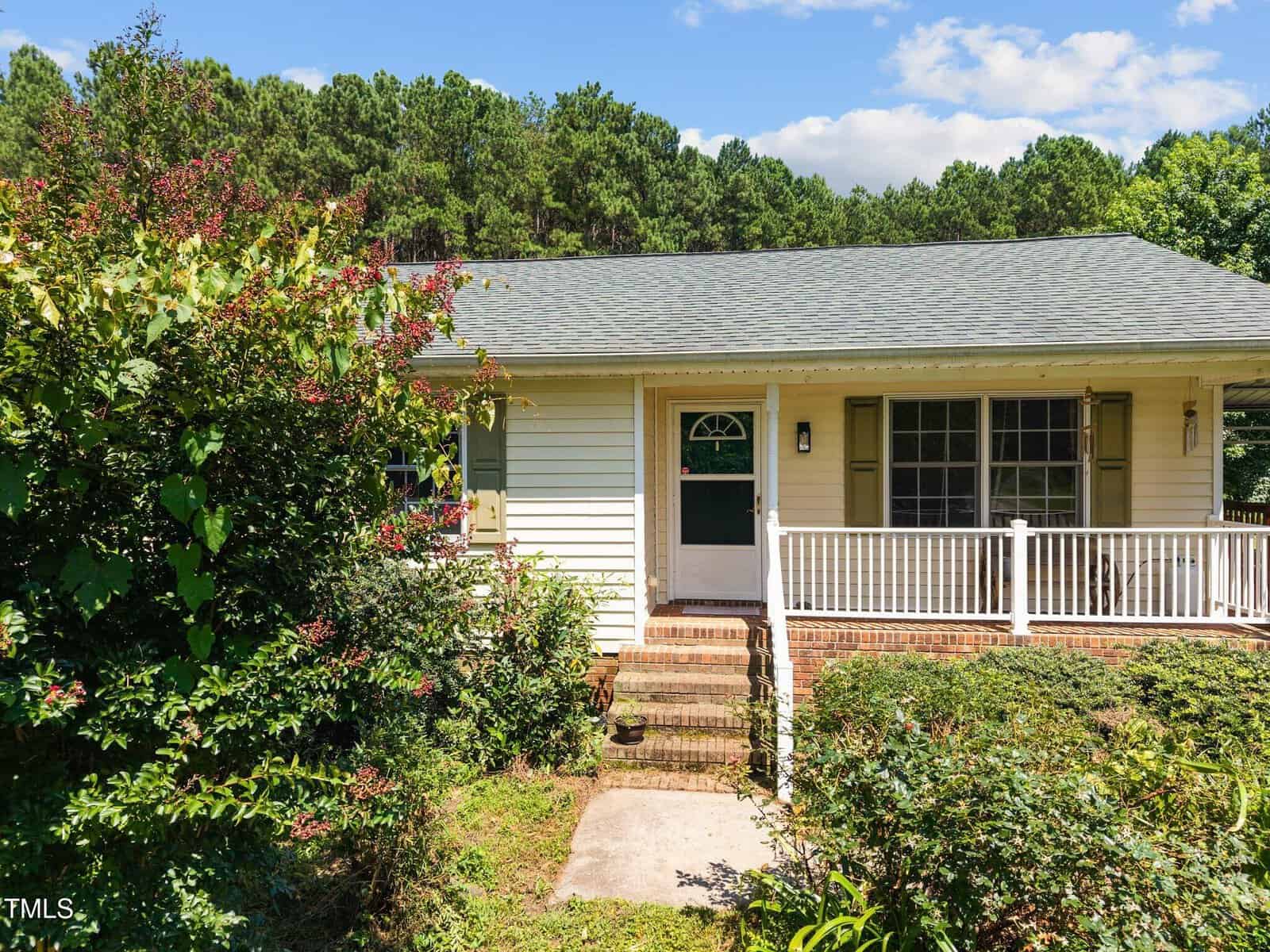 Single-story house with light-colored siding, a gray shingled roof, and a front porch with white railings, surrounded by lush green bushes and trees under a partly cloudy sky.