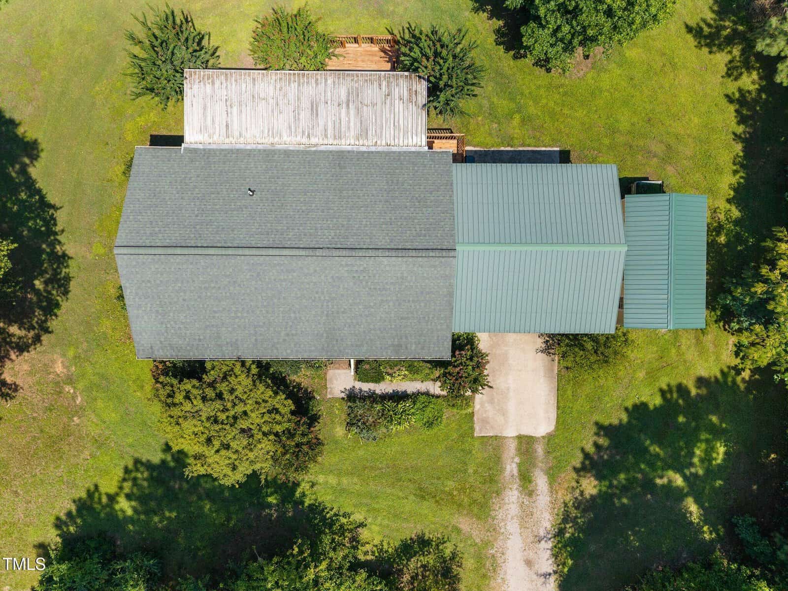 Aerial view of a house with a gray and green roof, a driveway, wooden deck, and surrounding green lawn and trees. The image is taken directly above, showing the structure and landscape layout.