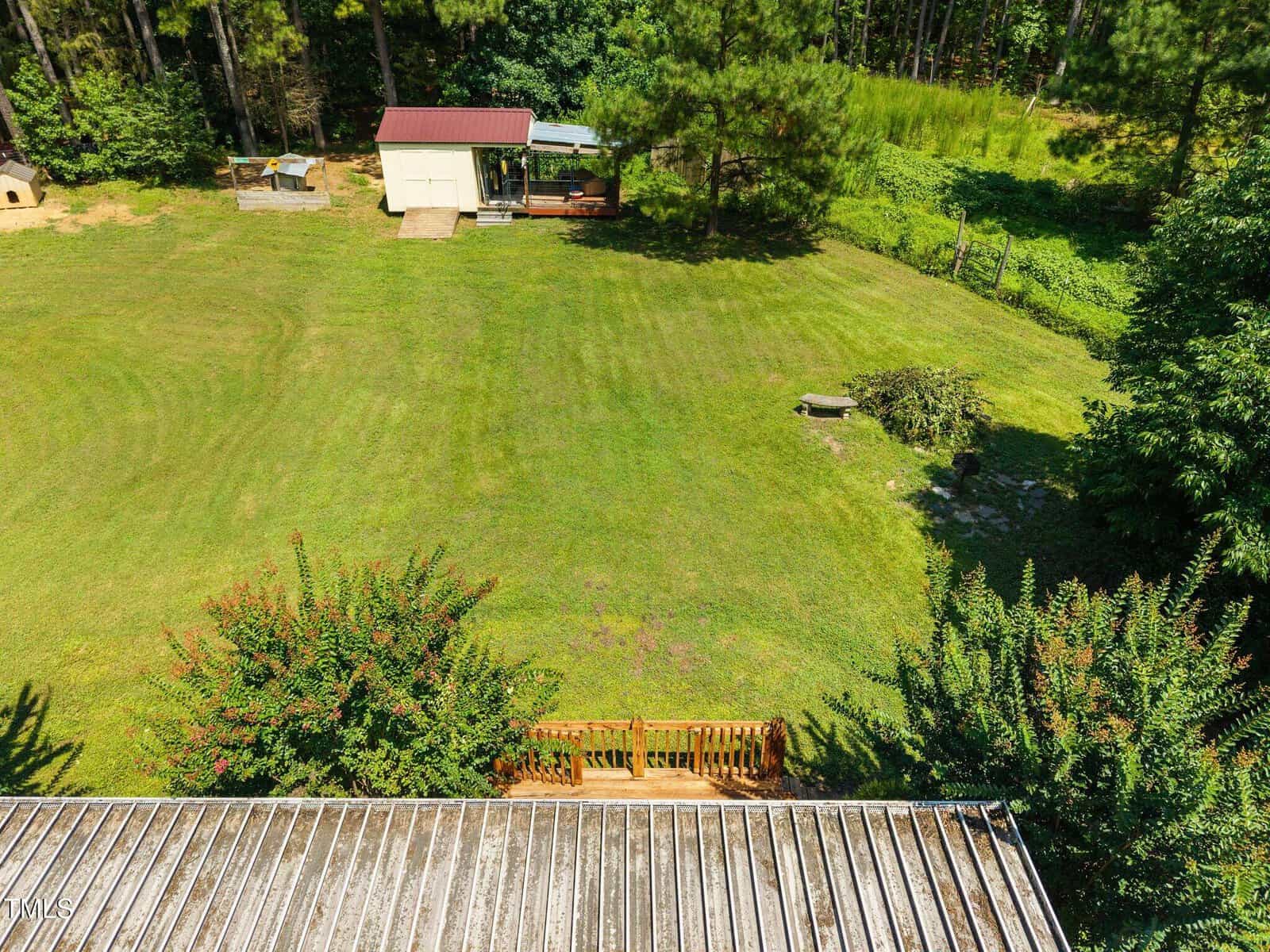 A large grassy backyard with a small shed, trees, and a wooden fence. There is a bench on the lawn and a doghouse near the fenced area. The roofing edge is visible at the bottom of the image.