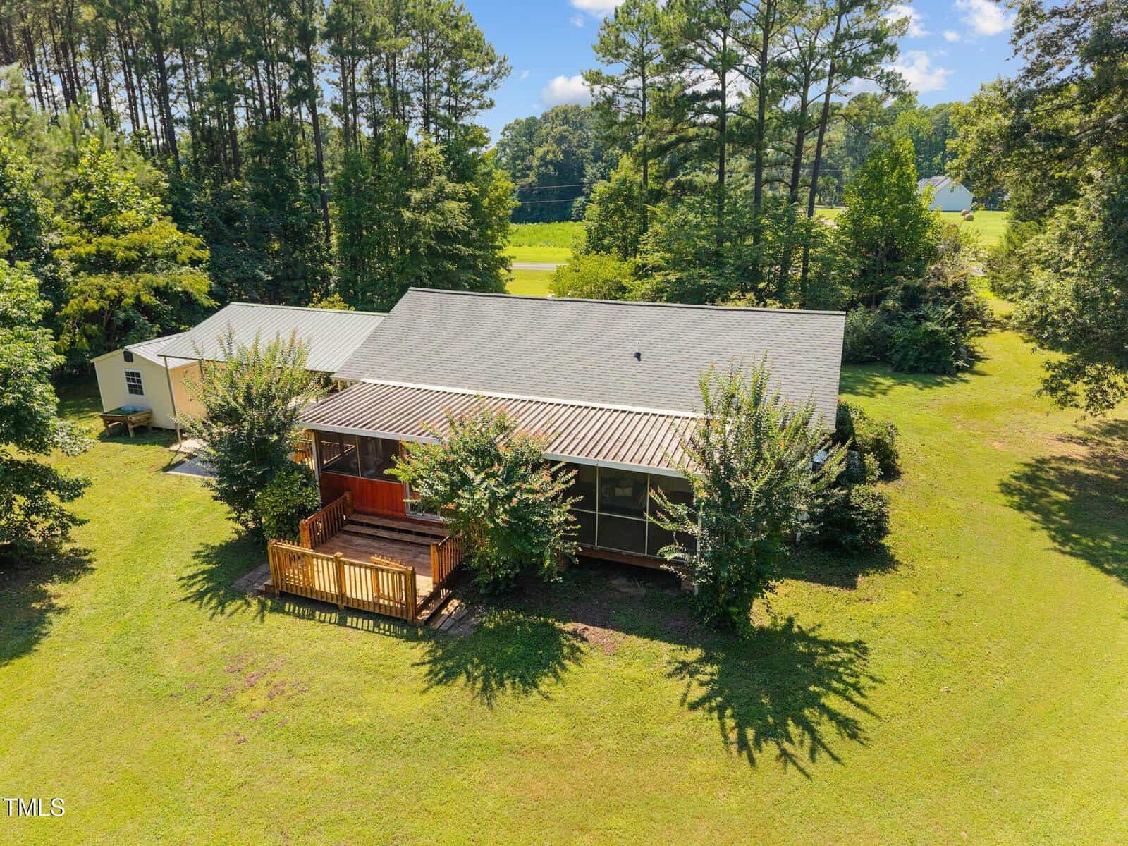 A single-story house with a screened porch and wooden deck sits on a grassy lot, surrounded by trees and bushes under a clear blue sky.