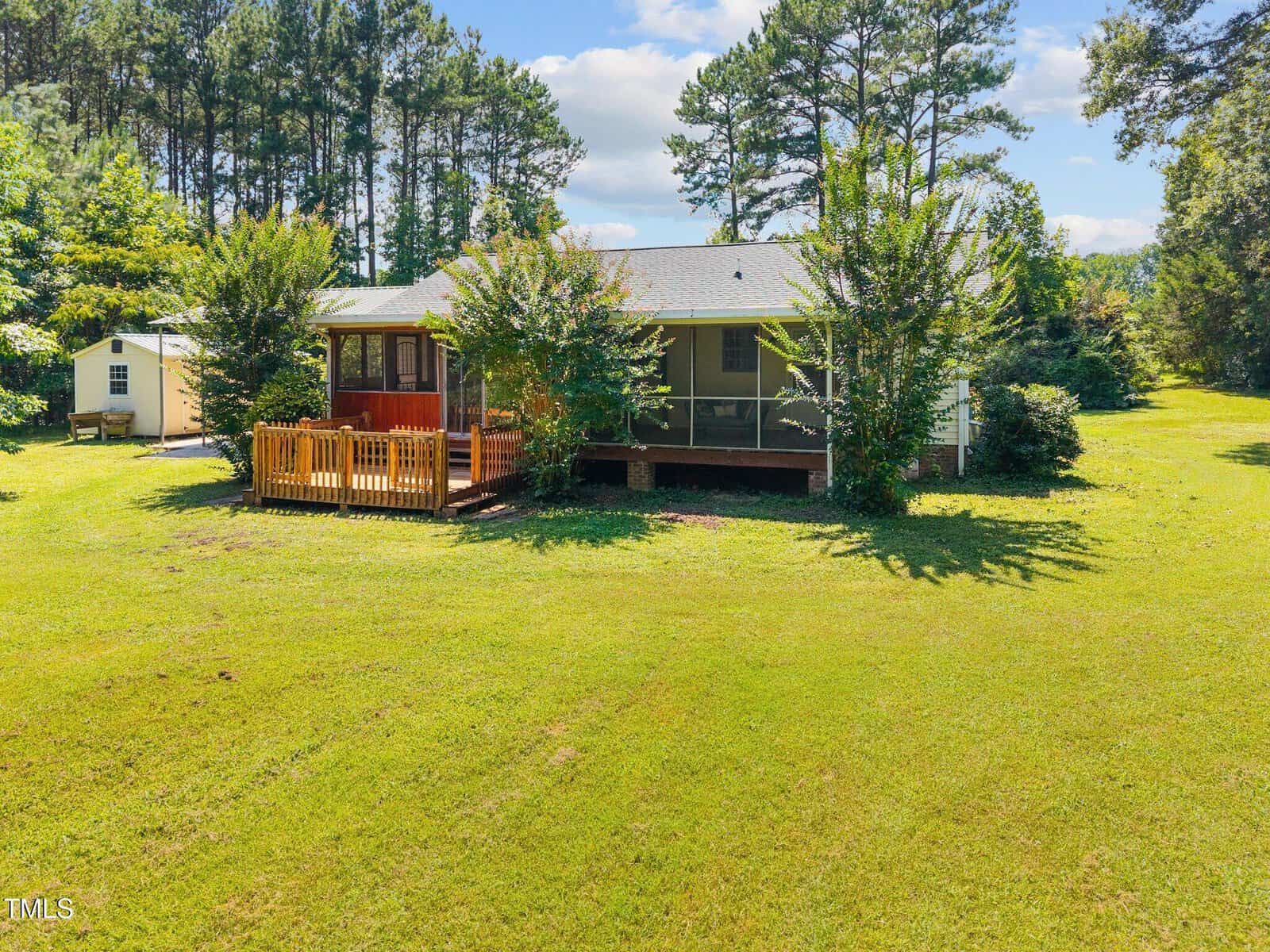 A one-story house with a screened porch and wooden deck sits on a large grassy yard surrounded by trees. A small shed is visible in the background. The sky is partly cloudy.
